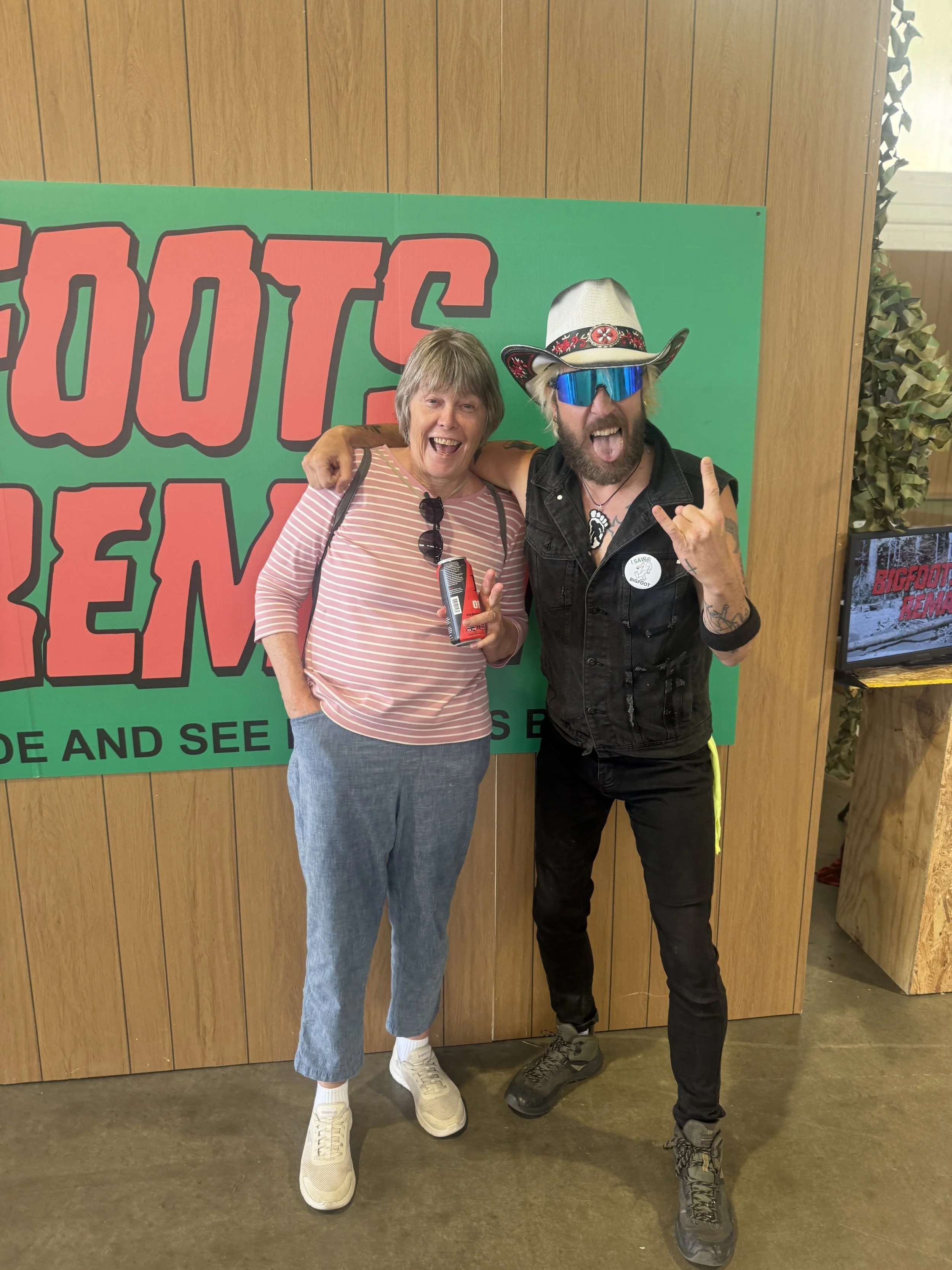 Two people posing together in front of a green poster with red text, one woman smiling and holding a can, and one man making a rock sign with his hand, wearing sunglasses and a cowboy hat.