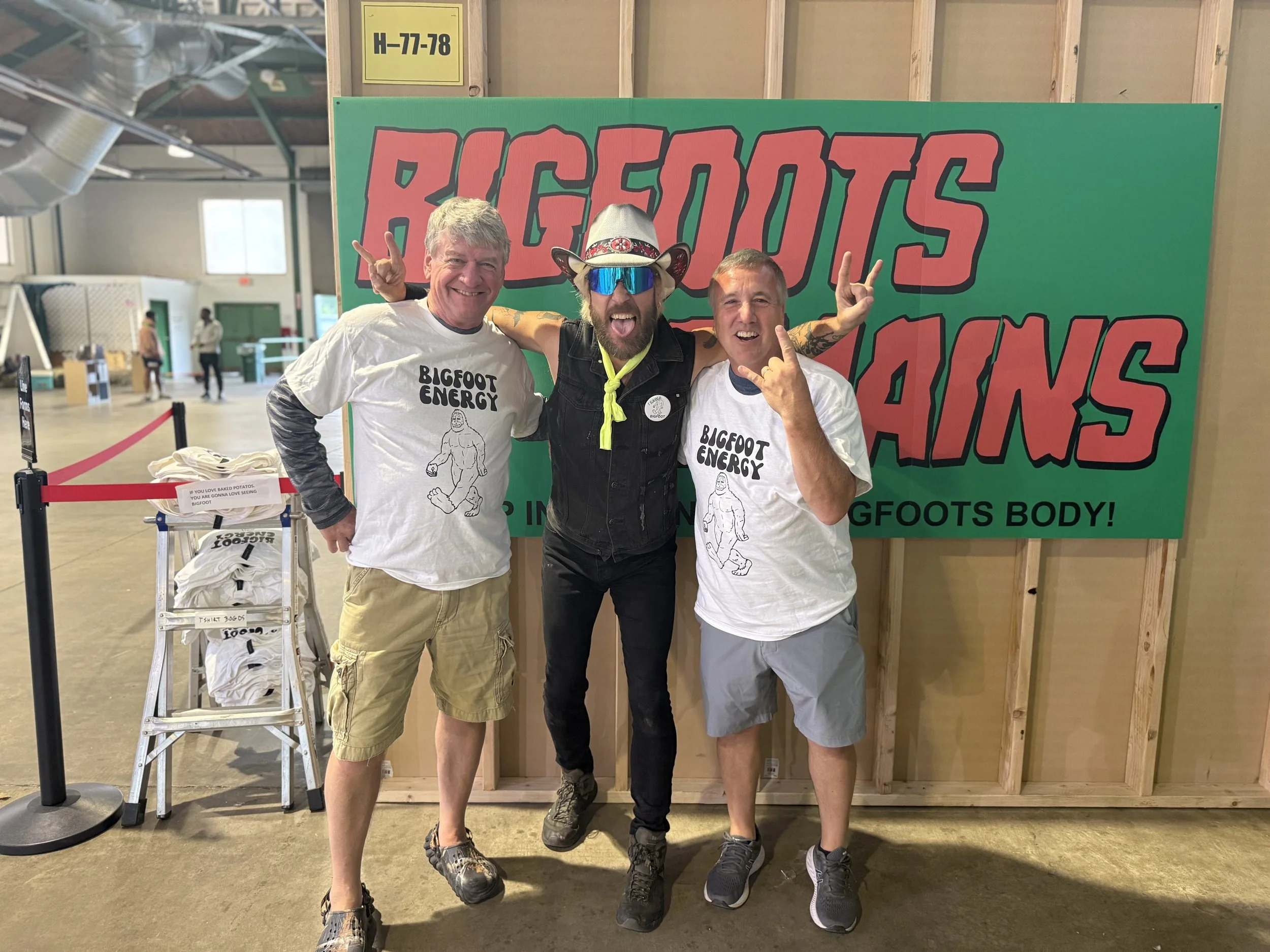 Three men standing in front of a sign that says 'BIGFOOTS BODY!' at a Bigfoot energy event, smiling, with the middle man wearing a cowboy hat and sunglasses, making a rock hand gesture.
