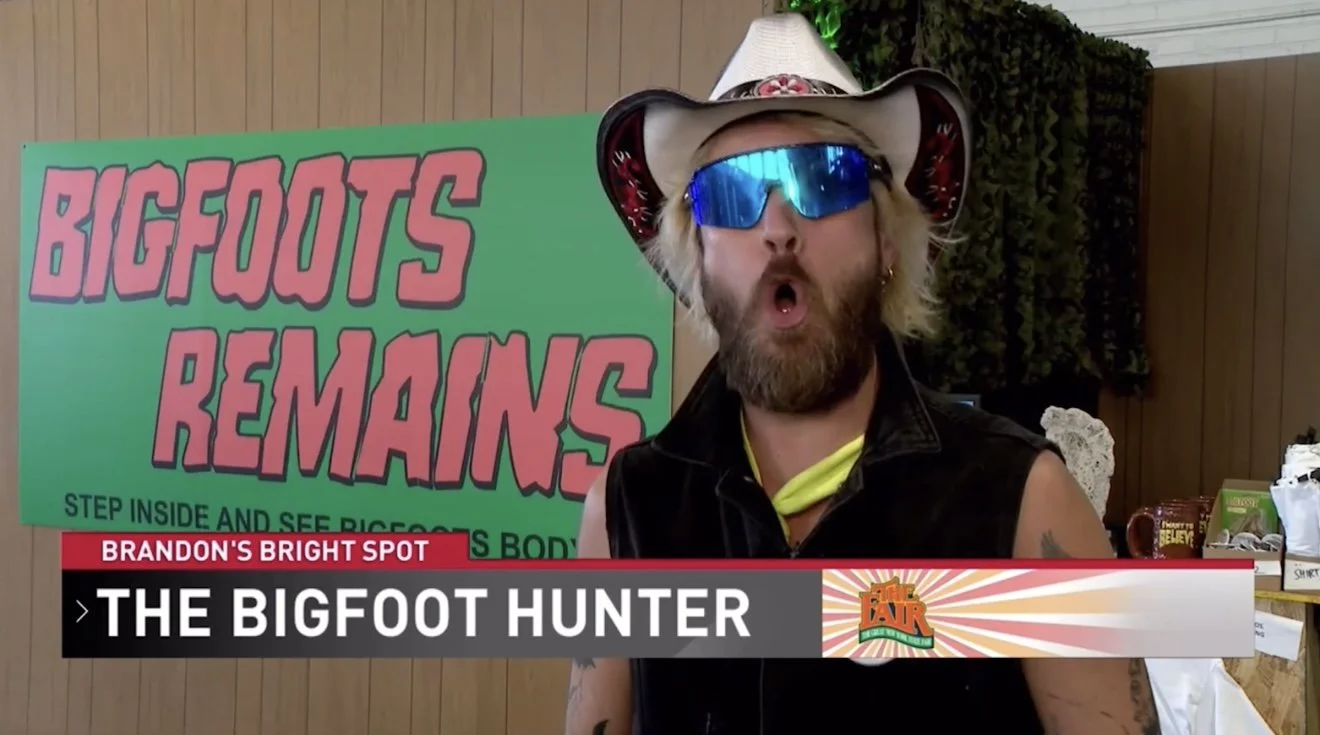 A man with a beard, wearing a large cowboy hat with a red band, reflective blue sunglasses, and a sleeveless black vest, is speaking to the camera. Behind him is a green sign with pink lettering that says "Bigfoot's Remains" and a smaller text that s
