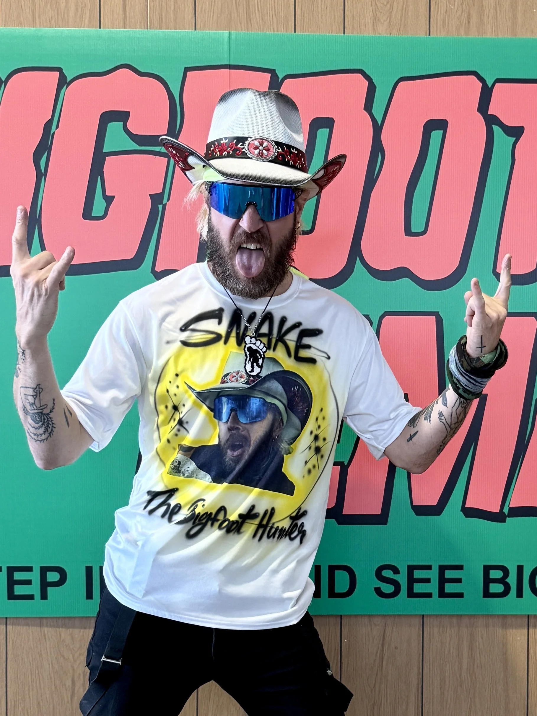 A man wearing a cowboy hat, reflective sunglasses, and a t-shirt with a graphic of himself. He is making a rock sign with both hands, sticking out his tongue, and posing in front of a colorful background with large cartoon-style text.