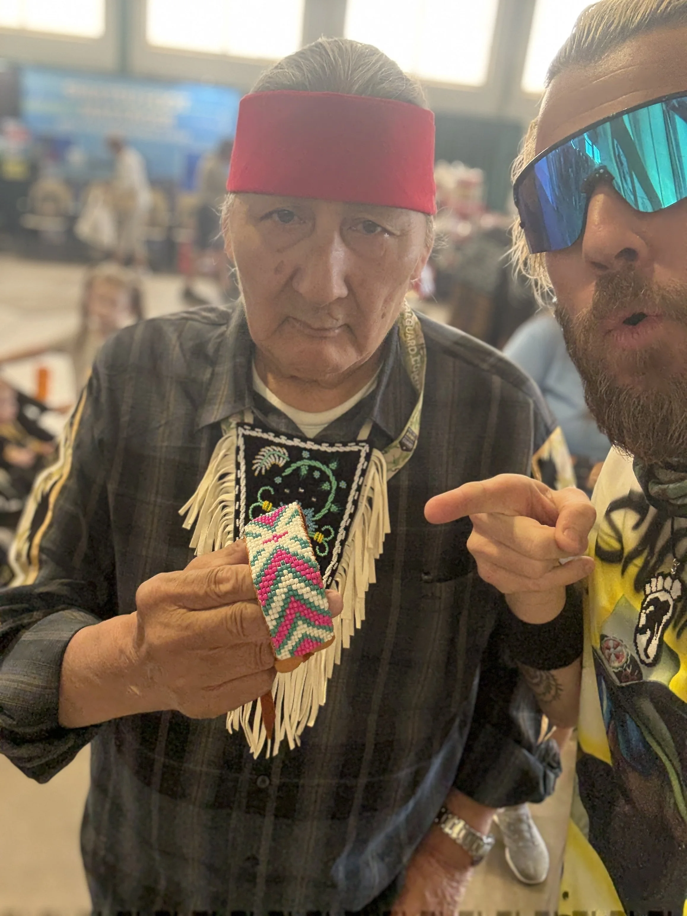 A man with long gray hair wearing a red headband and a plaid shirt holding a colorful beaded item, standing next to another man with sunglasses and a beard, in an indoor setting with blurred background.