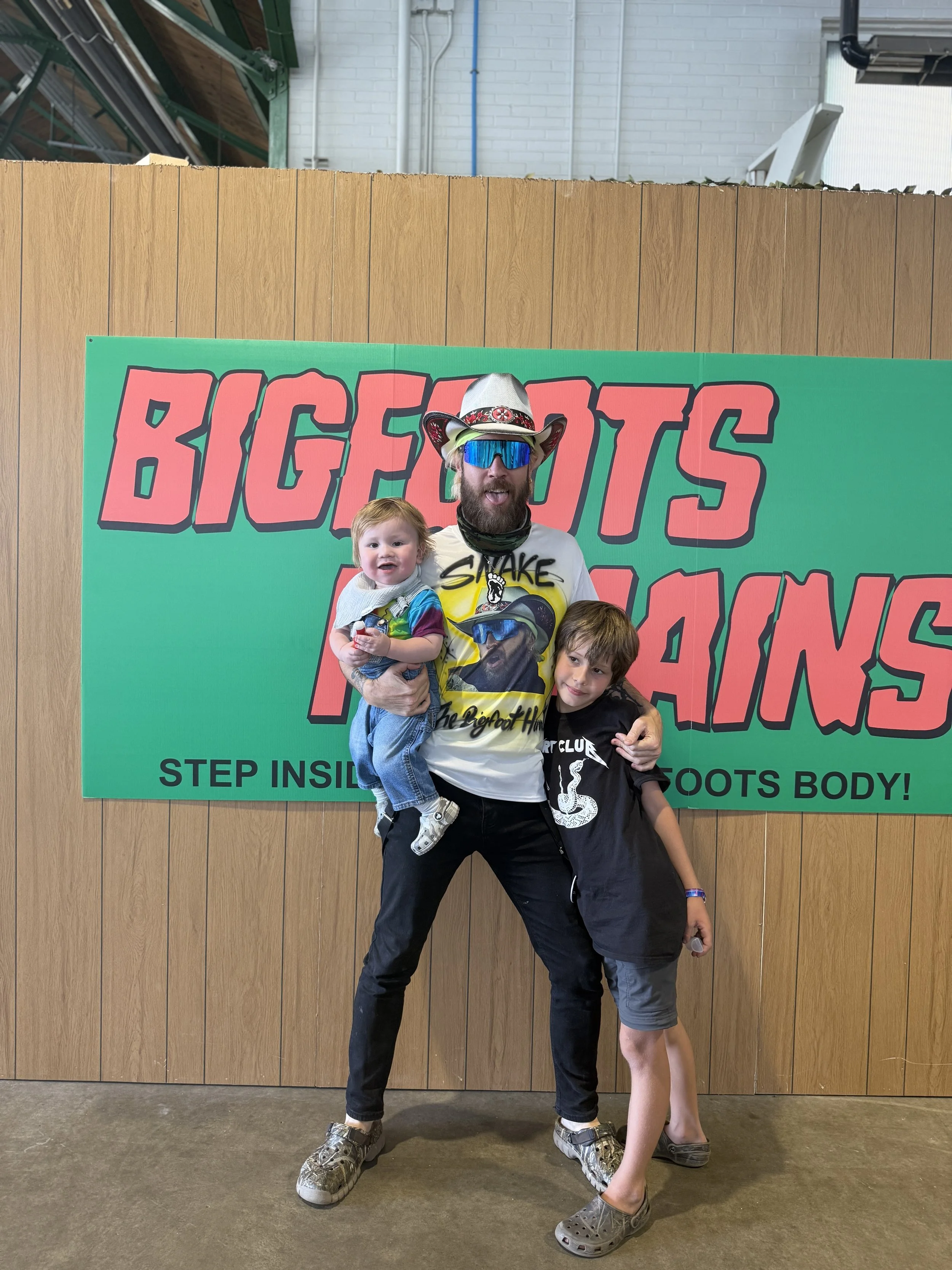 A man with a beard and sunglasses dressed as a cowboy, holding two children, standing in front of a sign that says "Bigfoot's Poots". One child is smiling and being held, the other is standing next to the man.