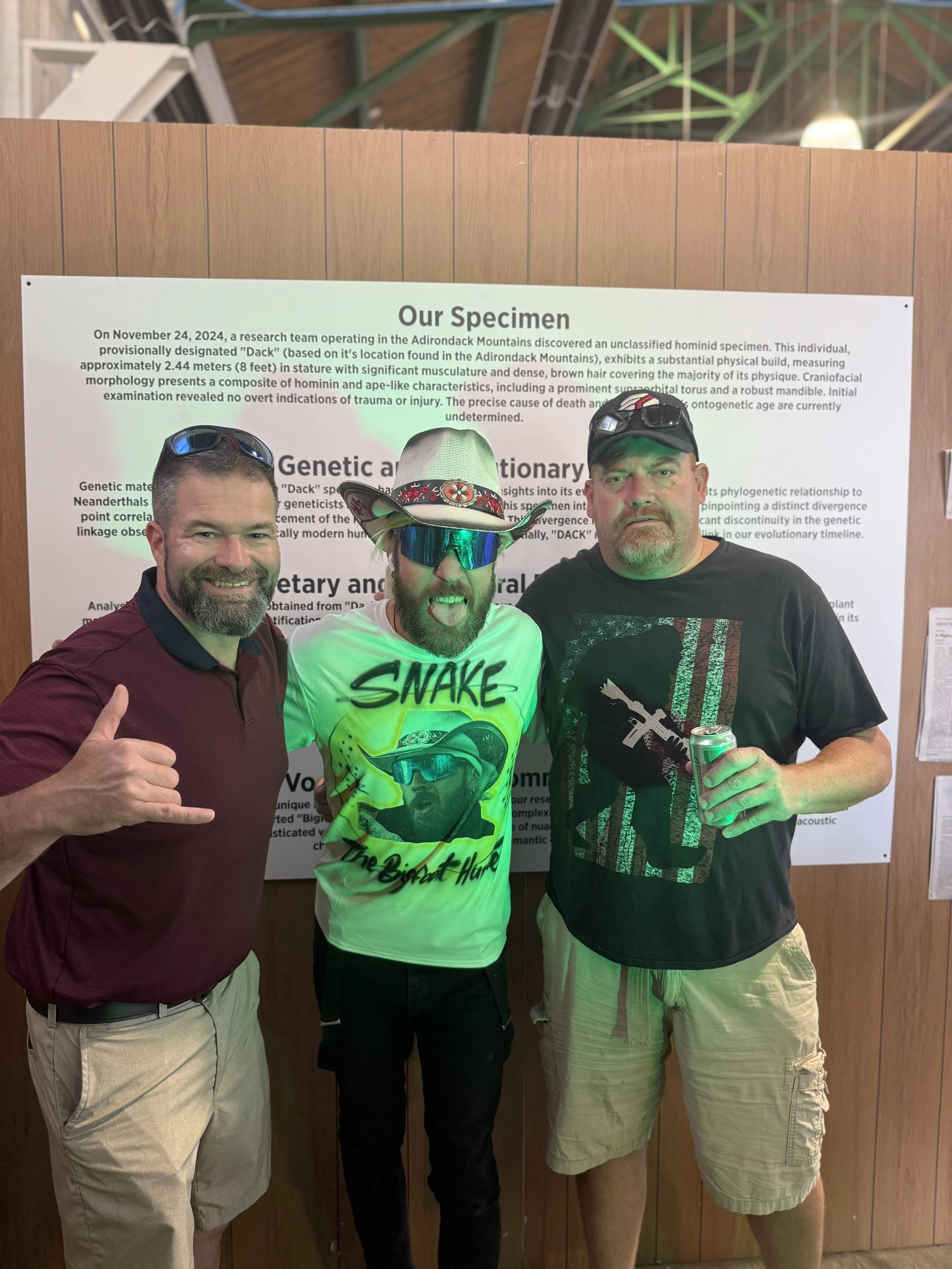Three men standing in front of a scientific poster at an indoor event, smiling at the camera. The man on the left is wearing a maroon polo shirt and khaki shorts, giving a shaka sign with his right hand. The man in the middle is wearing a graphic t-s