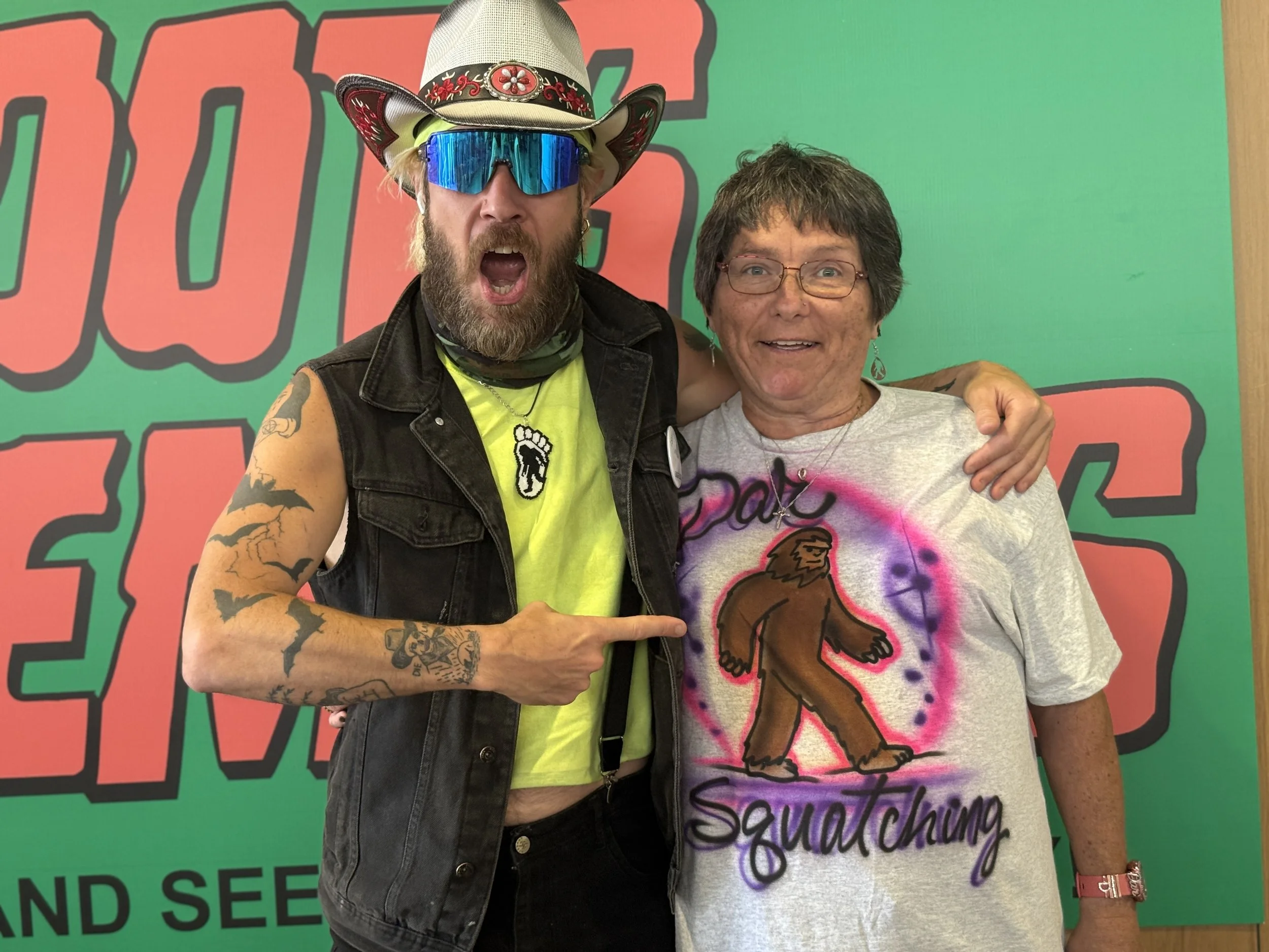 A bearded man wearing a cowboy hat, sunglasses, a yellow shirt, and a sleeveless denim vest, pointing at a woman with short gray hair and glasses. The woman is wearing a gray t-shirt with a printed, stylized Bigfoot figure and the words "Big Foot Squ