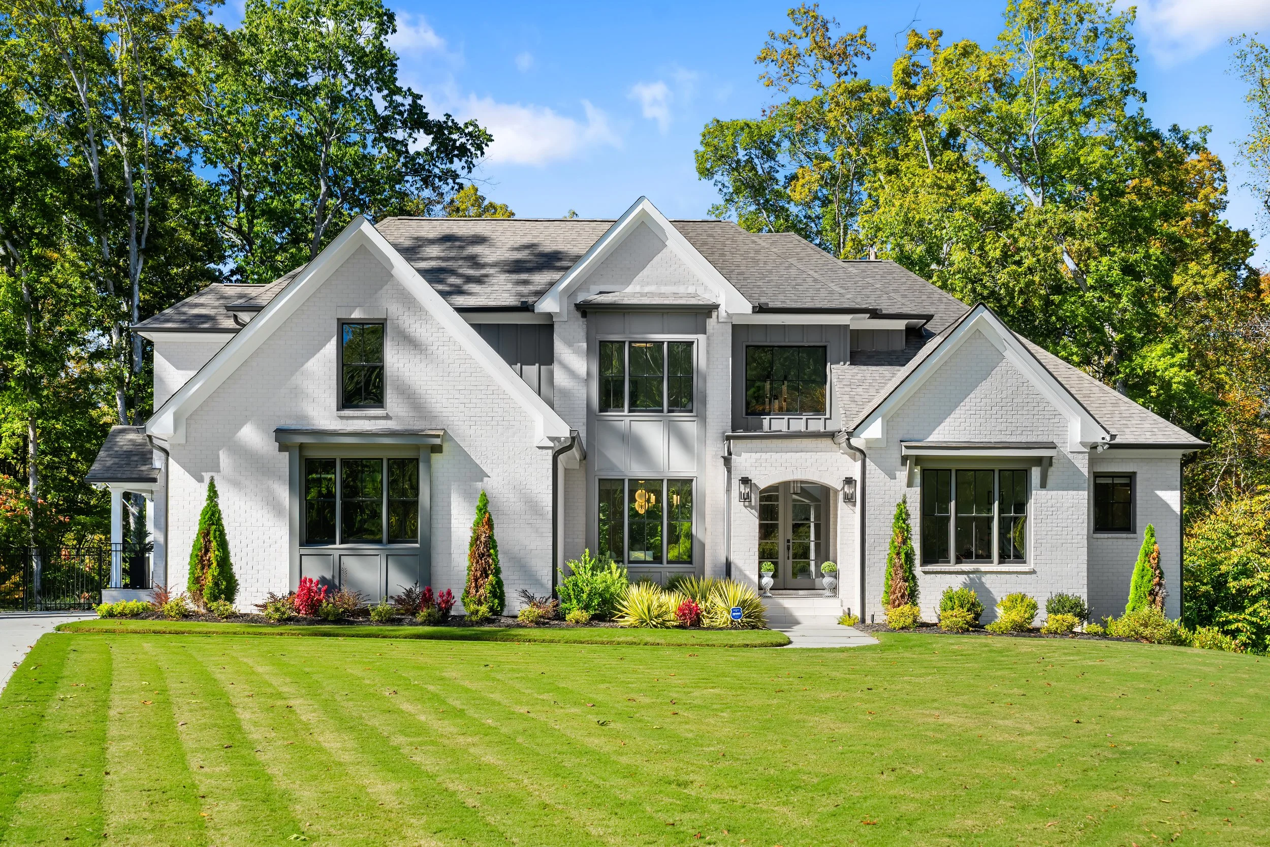 Large white two-story house with gabled roof and front porch, surrounded by a well-manicured lawn and trees under a blue sky.