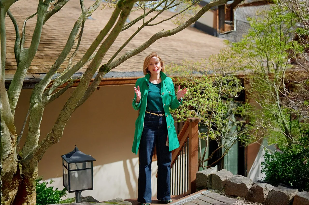 Smiling woman in blazer holding tablet outdoors with houses and plants in background