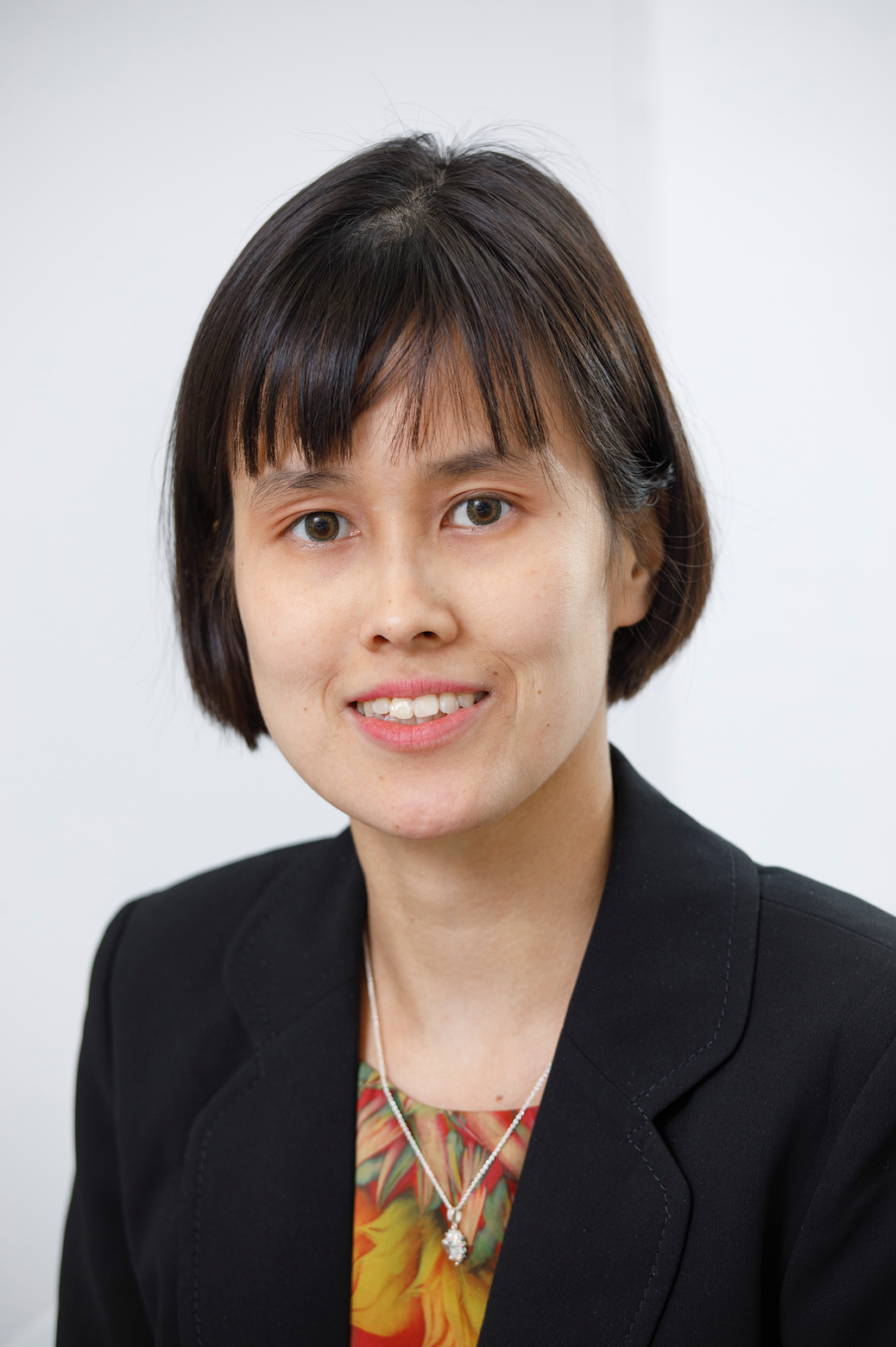 A woman with short dark hair, wearing a black blazer and a colorful blouse underneath, smiling at the camera against a plain white background.