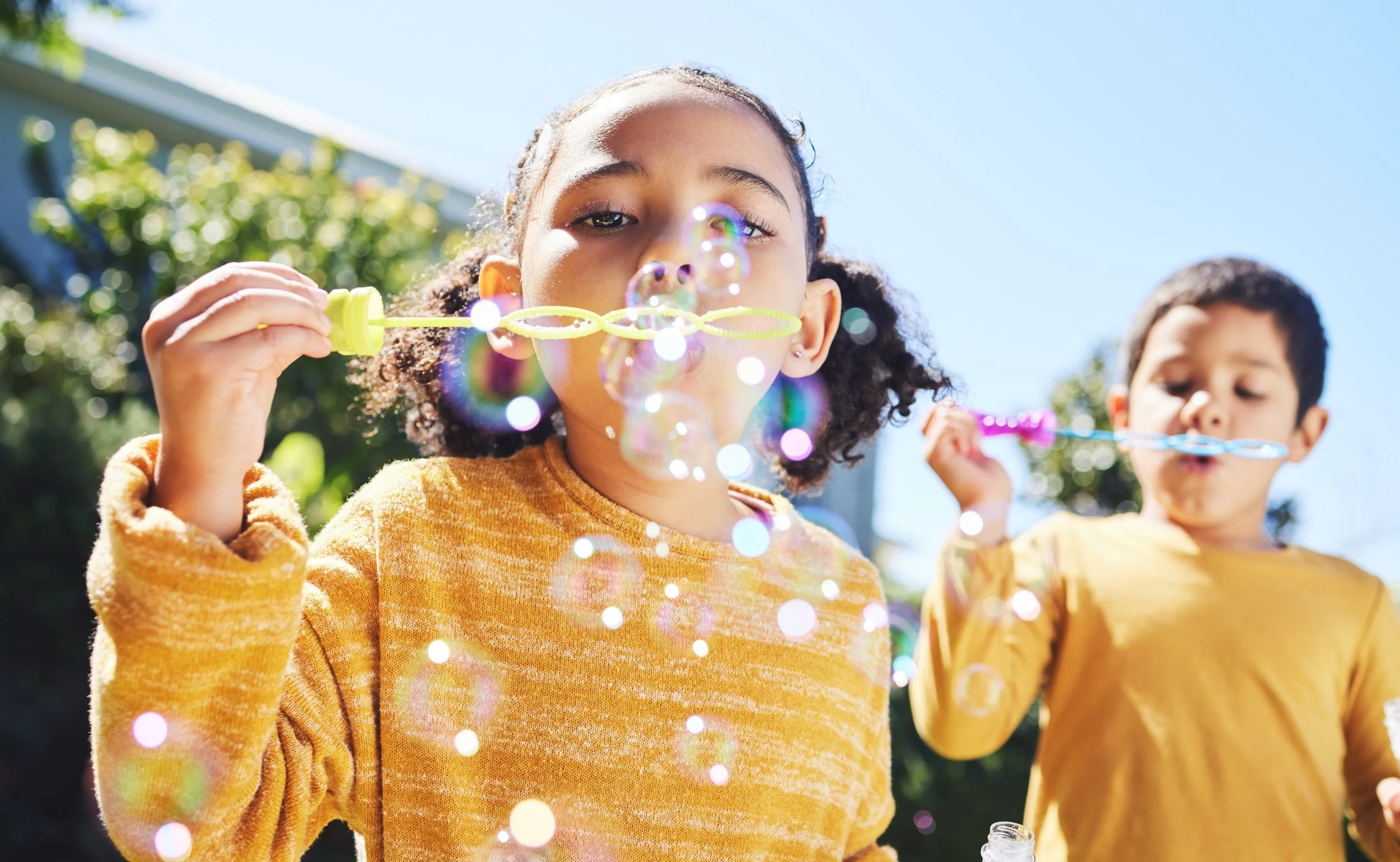 A young girl with curly hair blowing bubbles outdoors on a bright, sunny day, with another child in the background. She is wearing a yellow-orange sweater and is focused on blowing bubbles from a bubble wand.