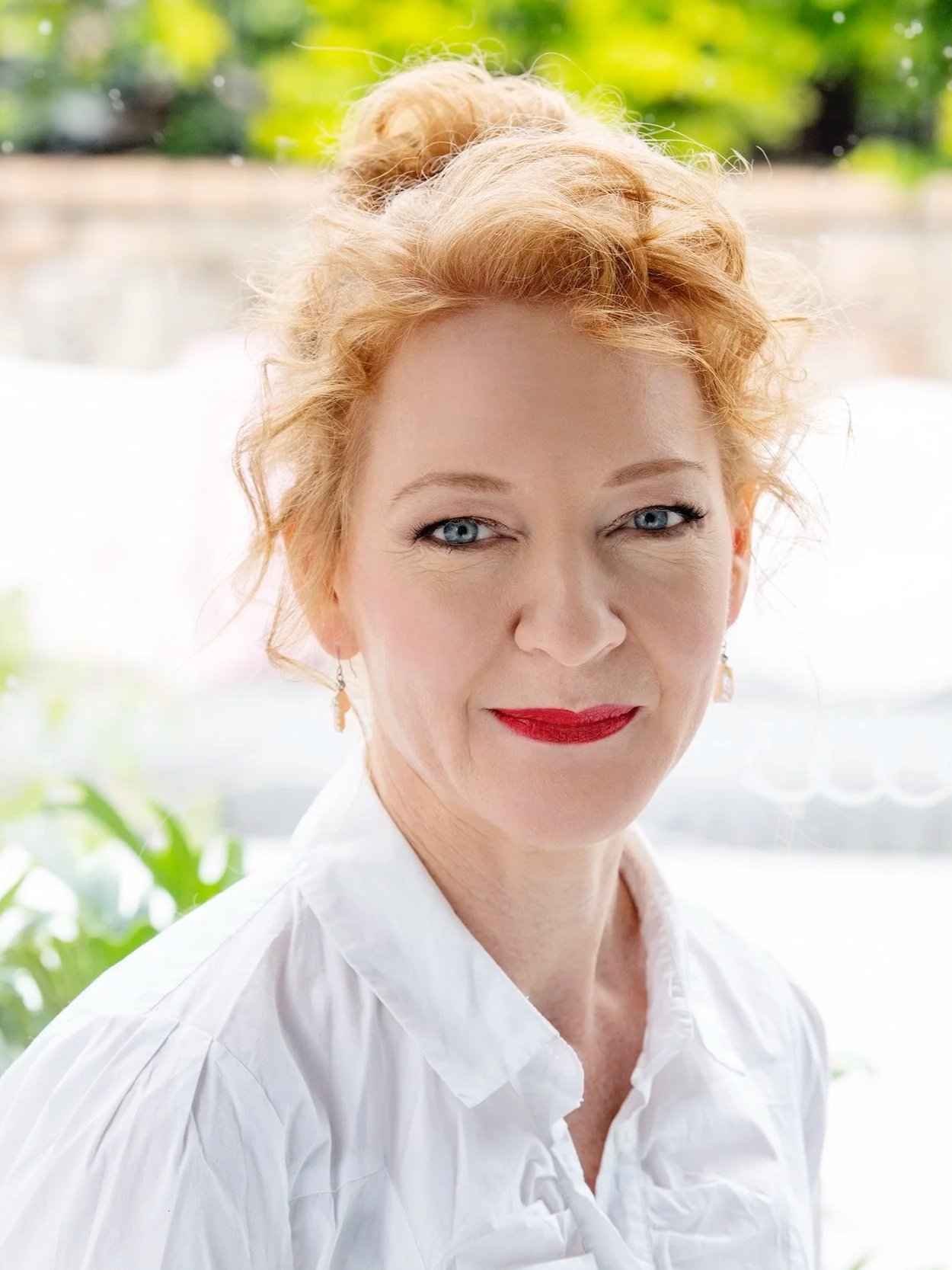 A woman with red curly hair, blue eyes, and red lipstick wearing a white blouse, sitting near a window with green foliage outside.
