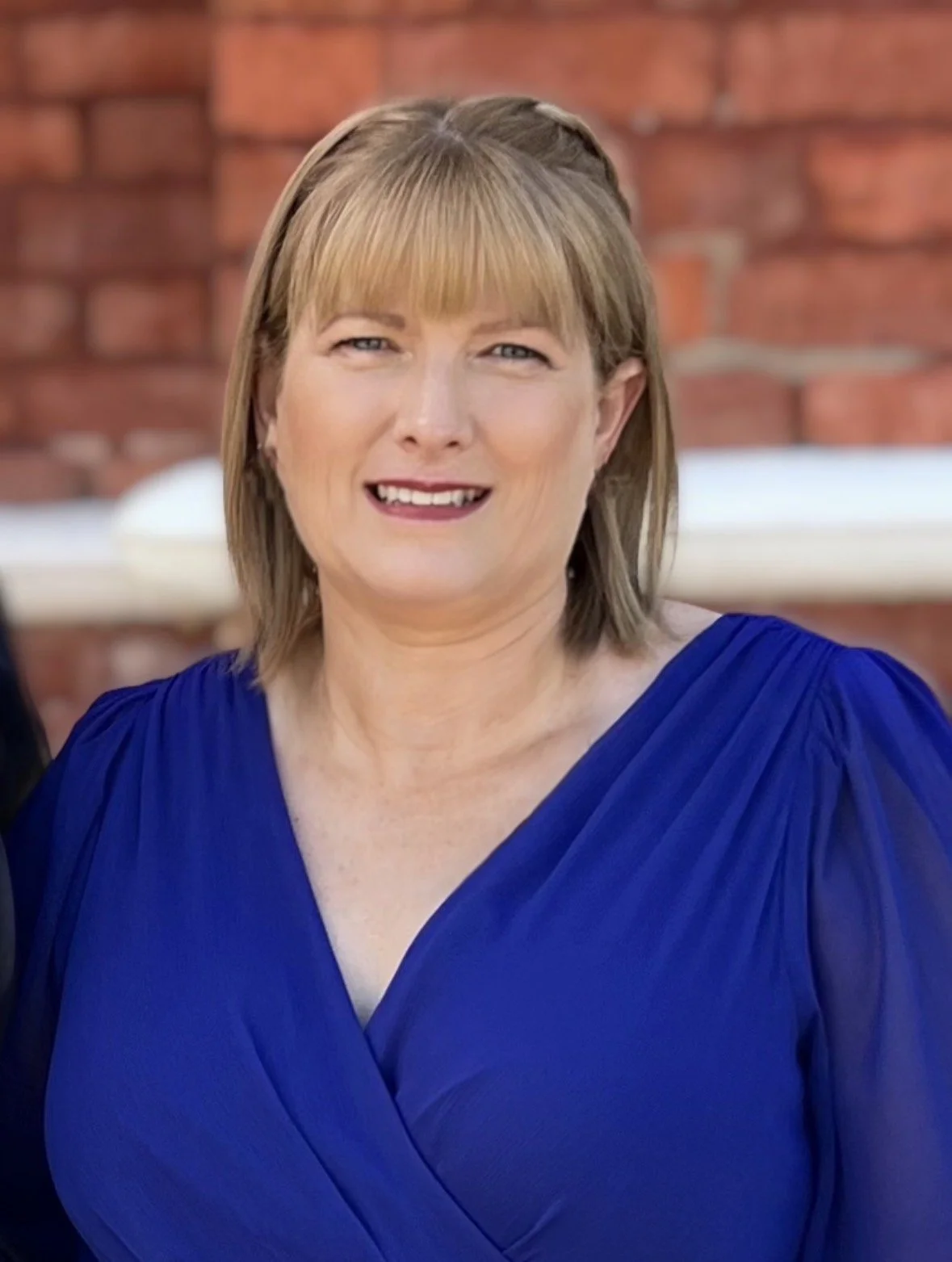 A woman with shoulder-length light brown hair and bangs, smiling, wearing a royal blue dress. She is outdoors with a brick wall in the background.