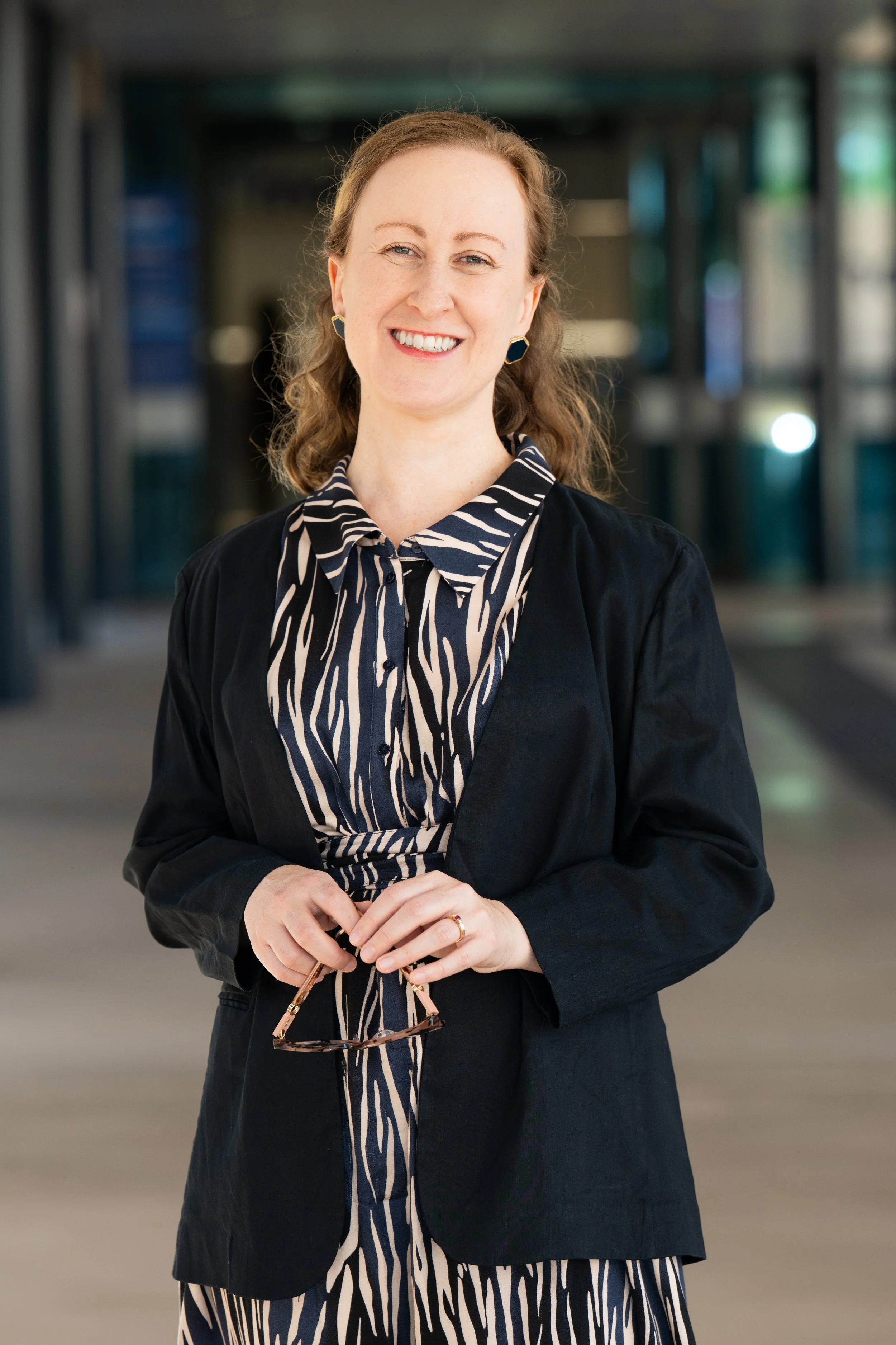 A woman with curly, shoulder-length hair smiling in an indoor setting, holding glasses, wearing a black blazer over a striped black and white dress with earrings.