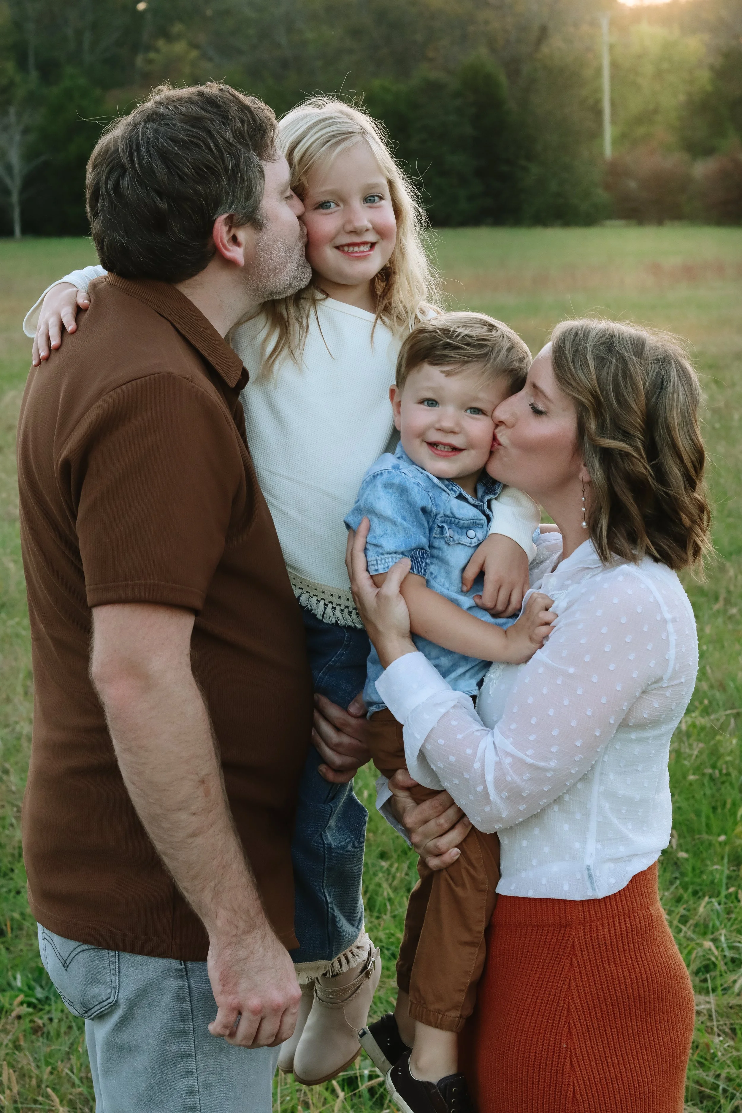 A family of four outdoors in a grassy field during sunset, a father giving a kiss on a daughter's cheek, a mother holding a young boy, all smiling and dressed casually.
