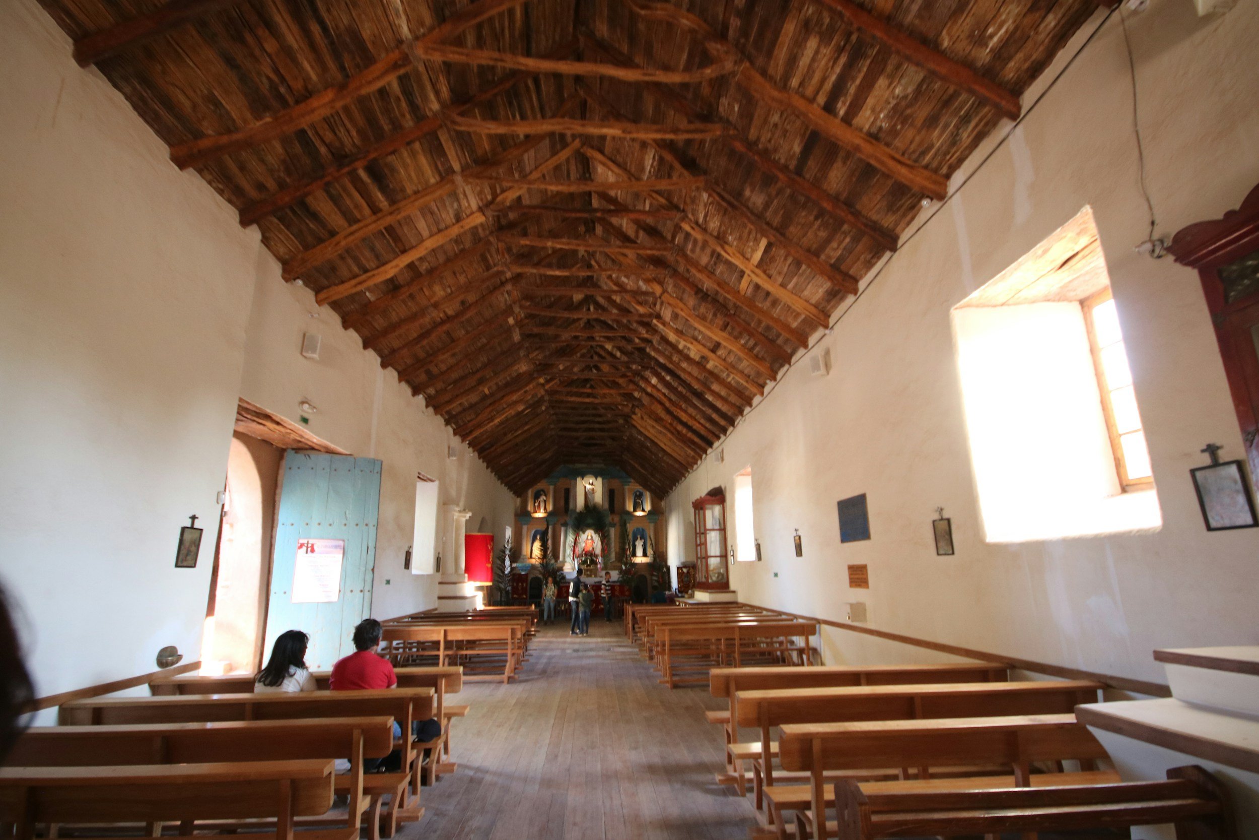 Interior of a church or chapel with wooden pews, a high wooden ceiling, and an altar at the far end. Churchgoers are seated on the pews, and sunlight streams through windows.