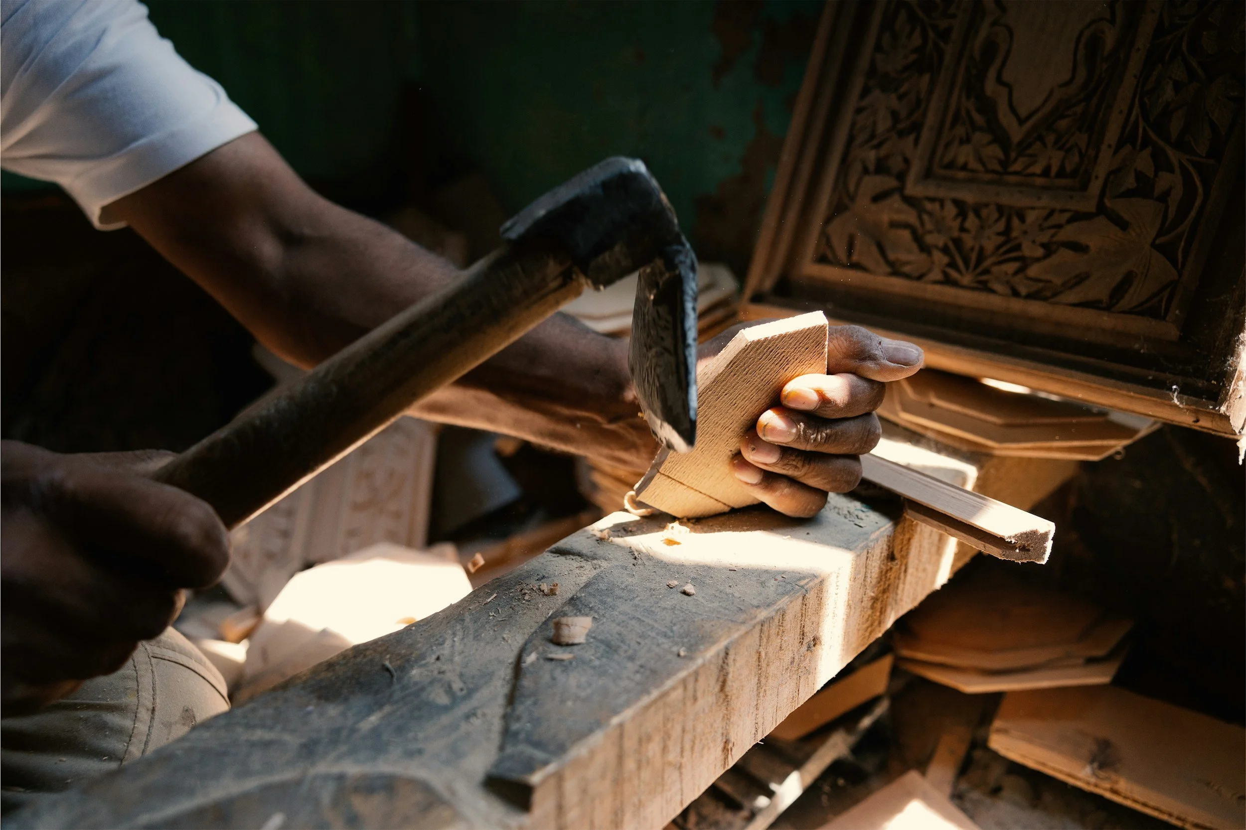 A person working on a woodworking project, holding a piece of wood with one hand and using a hammer with the other, with carved wooden panels in the background.