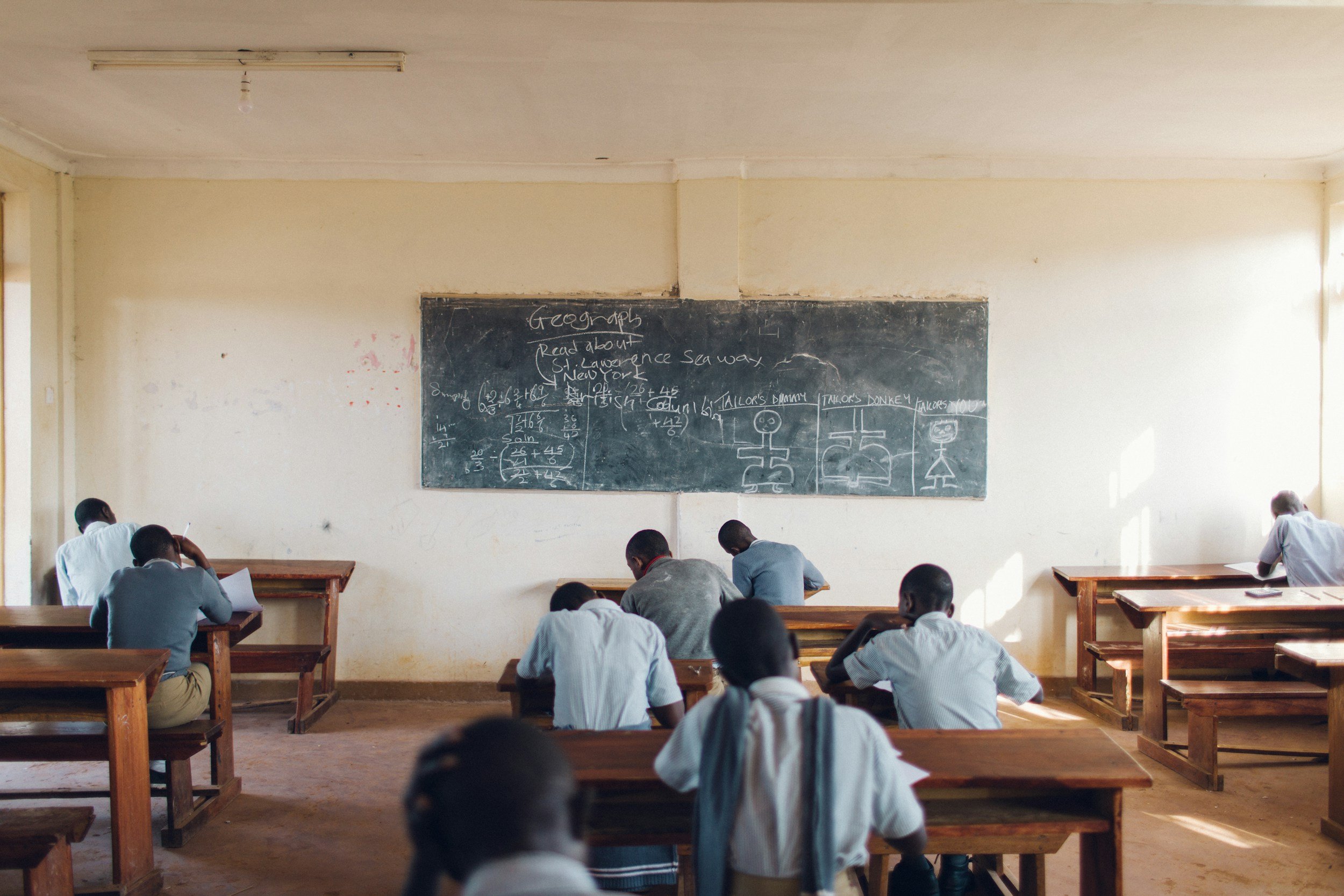 A classroom with students seated at wooden desks, facing a chalkboard with math equations and drawings, in a room with bright natural light.