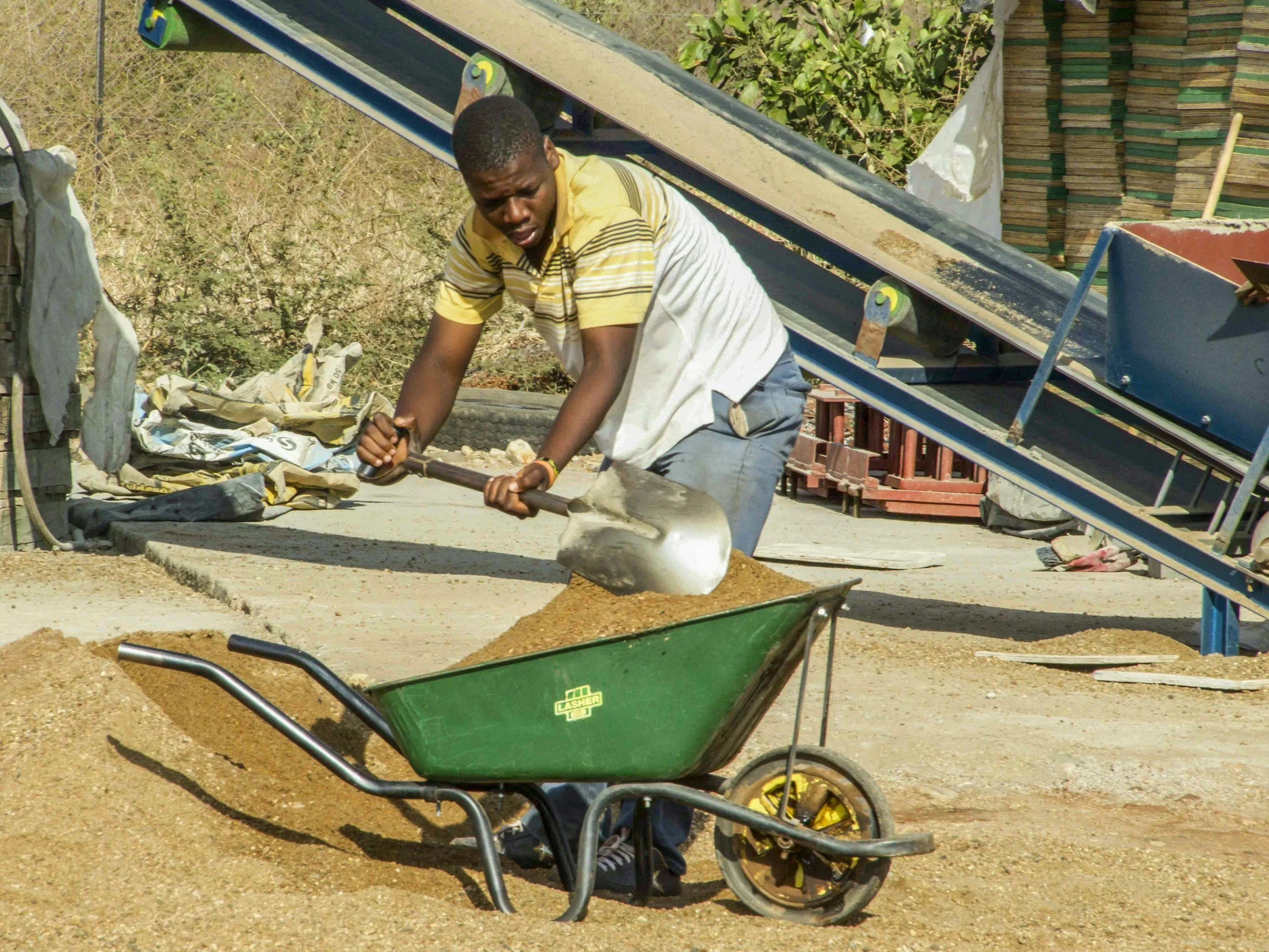 Young man working on a concrete slab with a shovel, with a wheelbarrow in the foreground, and construction materials and greenery in the background.