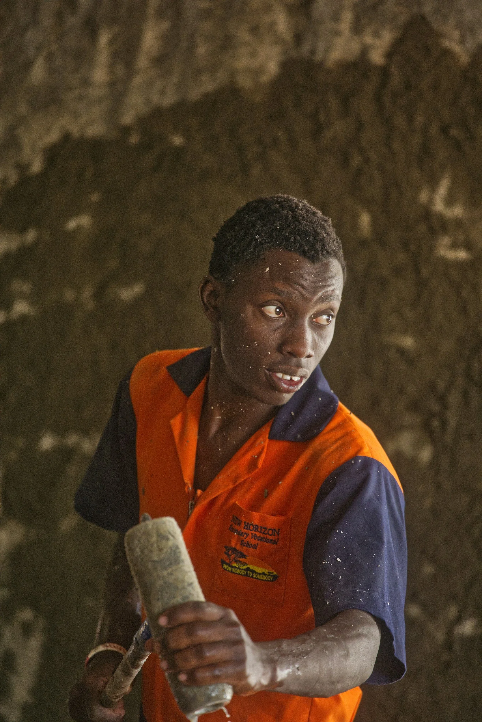 A young man with short curly hair in an orange and navy blue shirt holding a rock climbing tool or stick, with a blurred natural rock wall or cave as background.