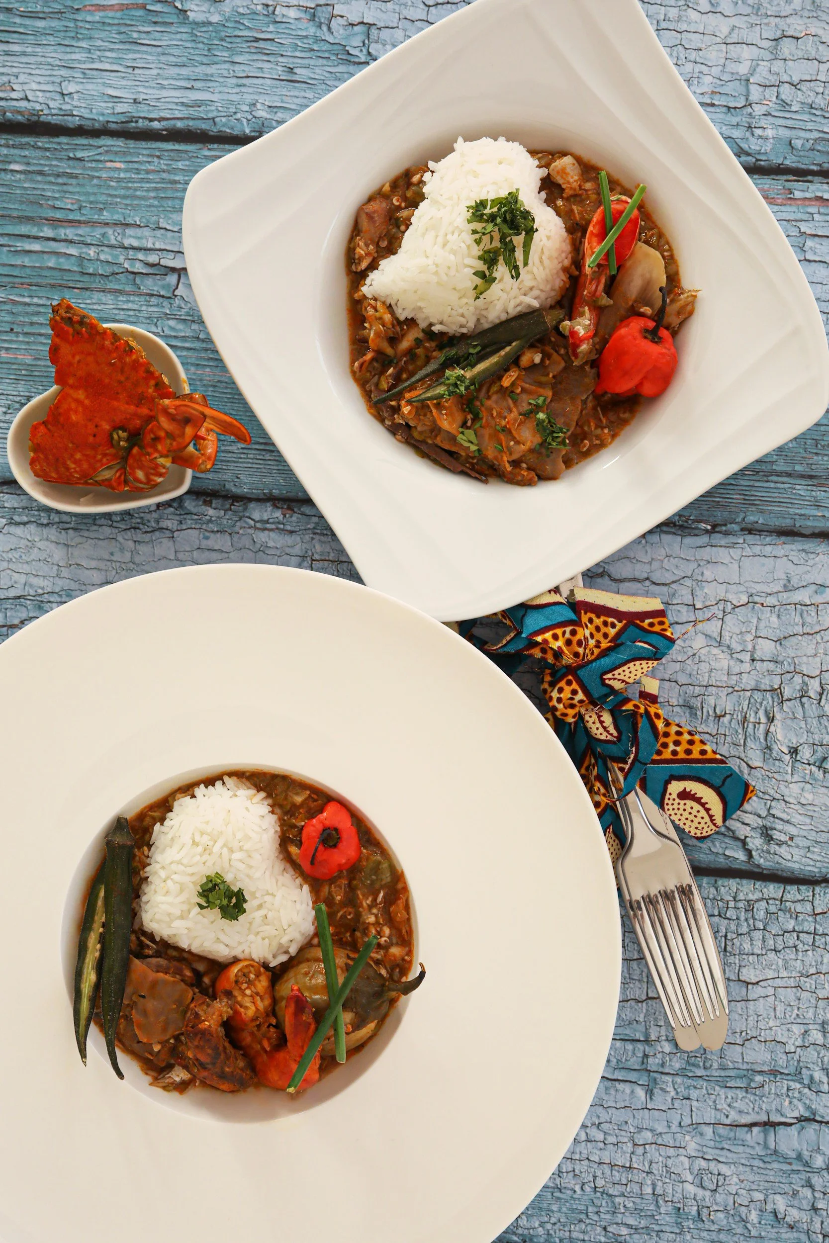 Two plates of rice with curry, vegetables, and garnishes on a blue, textured wooden table, with a silver fork and a colorful cloth napkin.