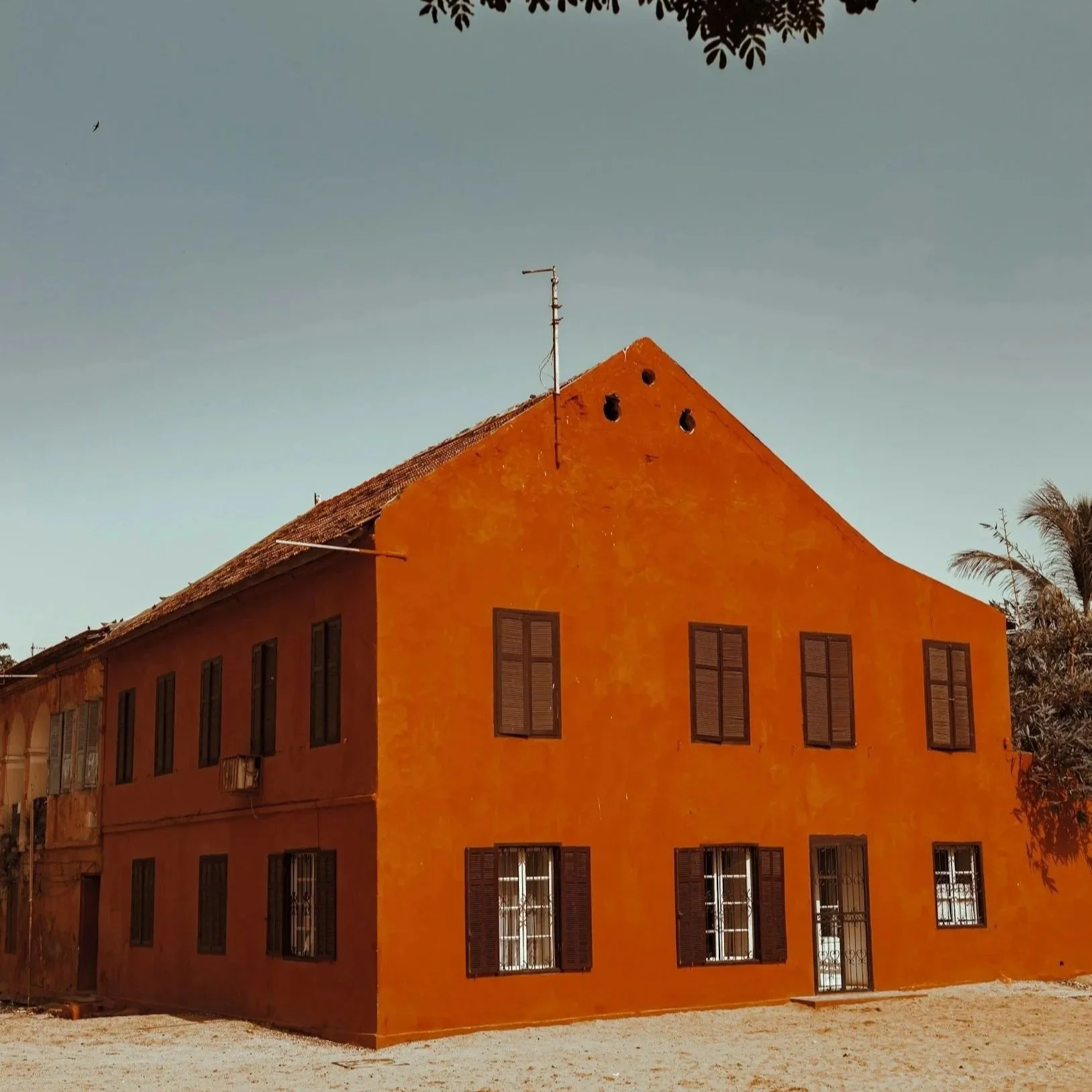 An orange-colored building with multiple tall, rectangular windows and a sloped roof. There are trees and a clear sky in the background.