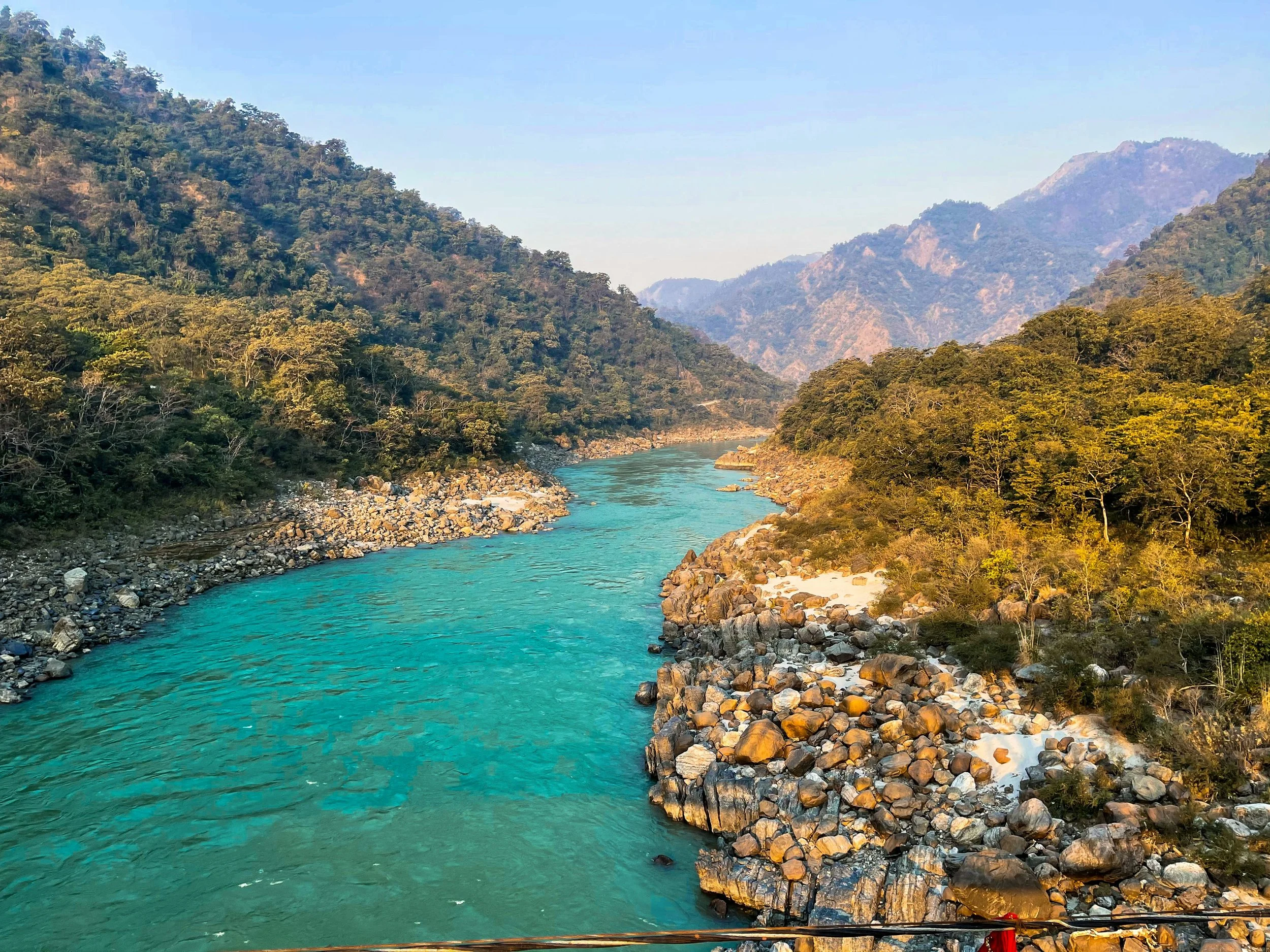 The Gangees River in Rishikesh, India flowing through a forested valley with mountains in the background and rocks along the shoreline.