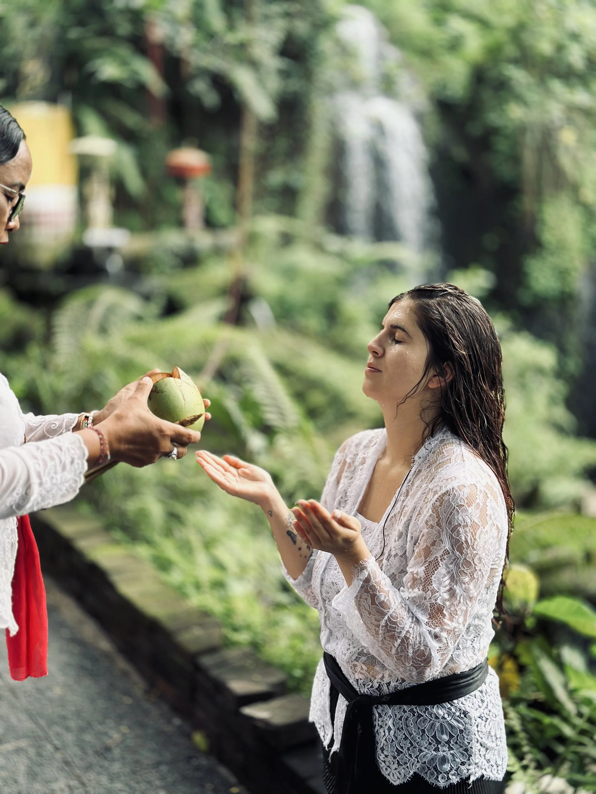 A woman in a white lace top with a black belt, standing outdoors with lush greenery and waterfalls in the background. She is receiving blessings and purification in a traditional Balinese waterfall cleansing ceremony.