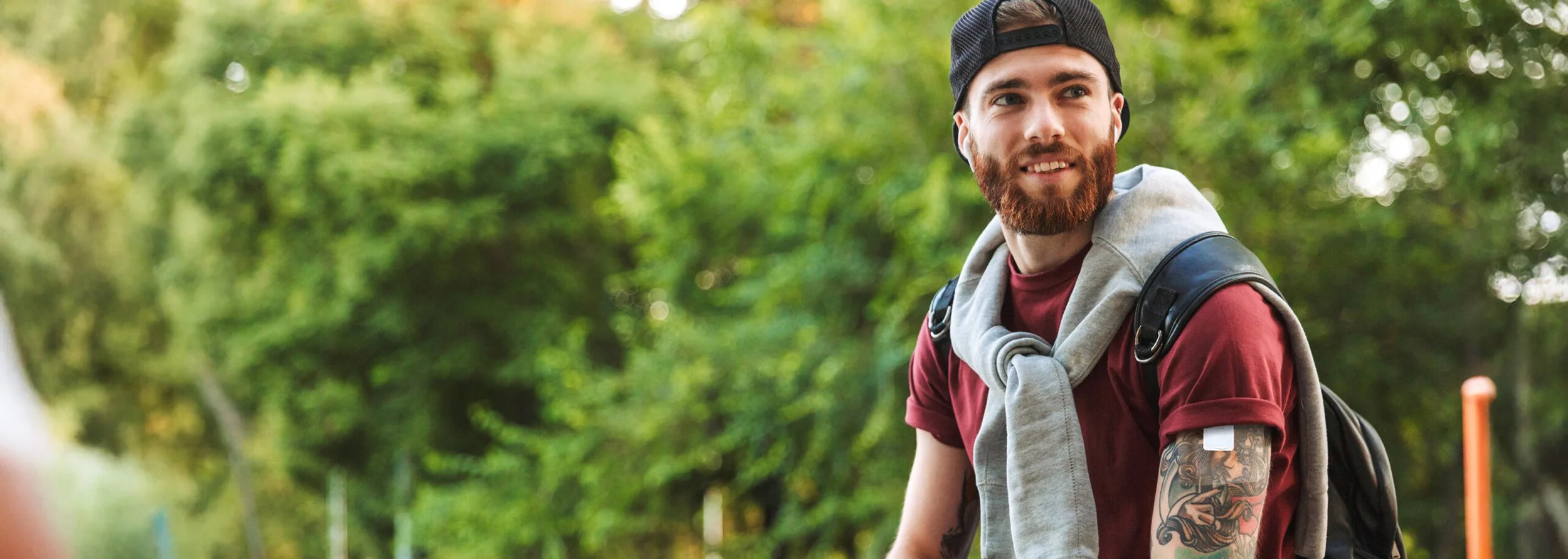 A young man with a beard, wearing a backwards baseball cap, red shirt, and gray sweatshirt tied around his shoulders, smiling outdoors with green trees in the background, carrying a backpack.