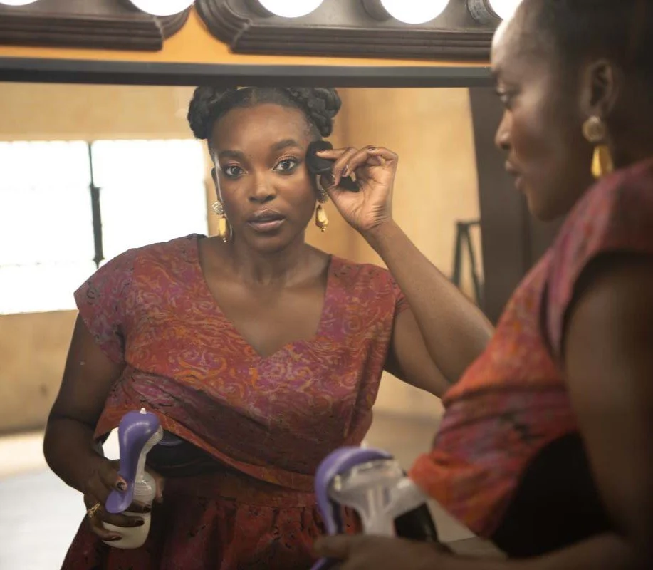 Wunmi wearing a red patterned Iyadé maternity dress applies makeup while looking into a mirror.