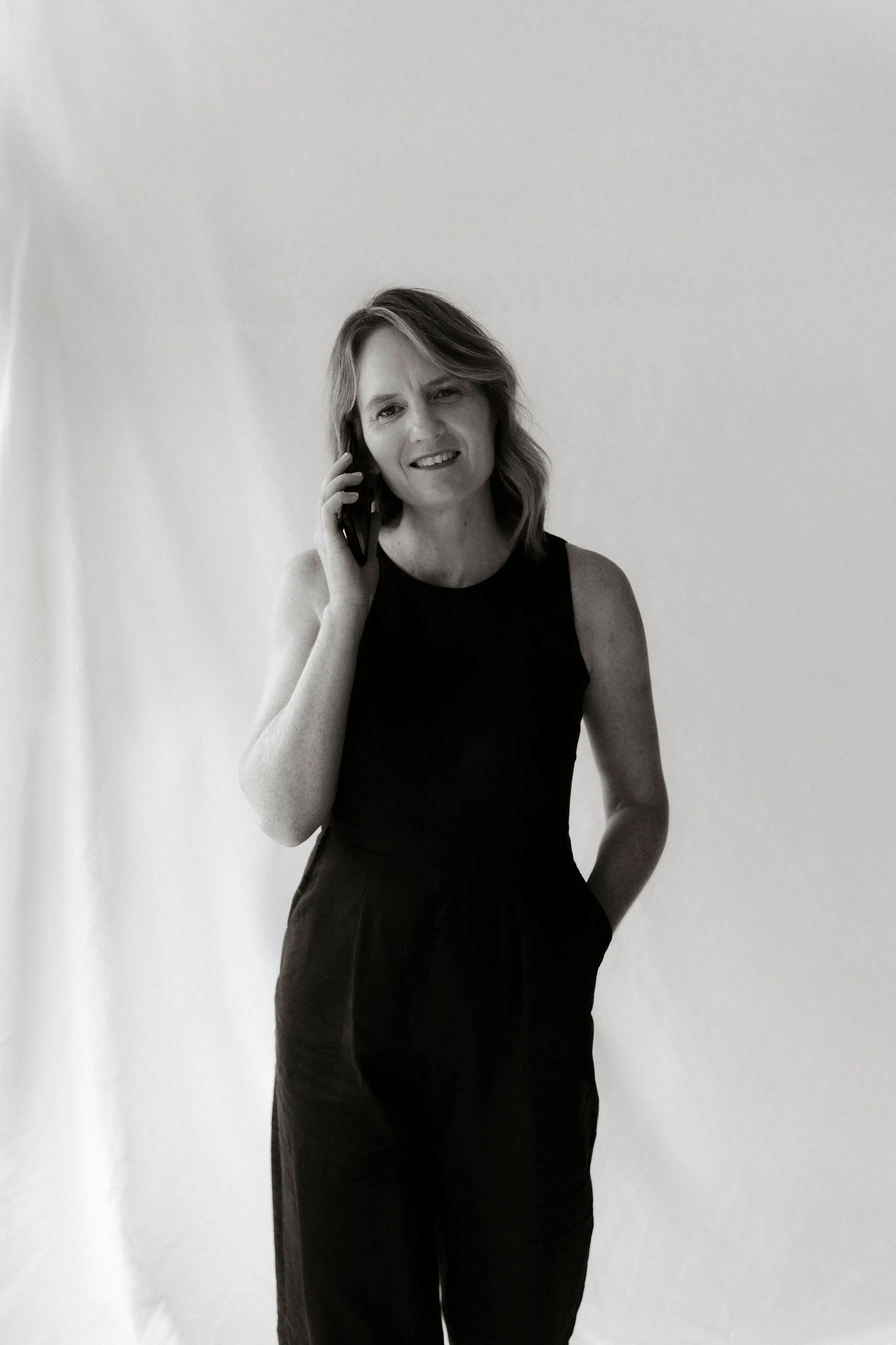 Black and white photo of a person using a smartphone, wearing a sleeveless outfit, standing against a plain background.