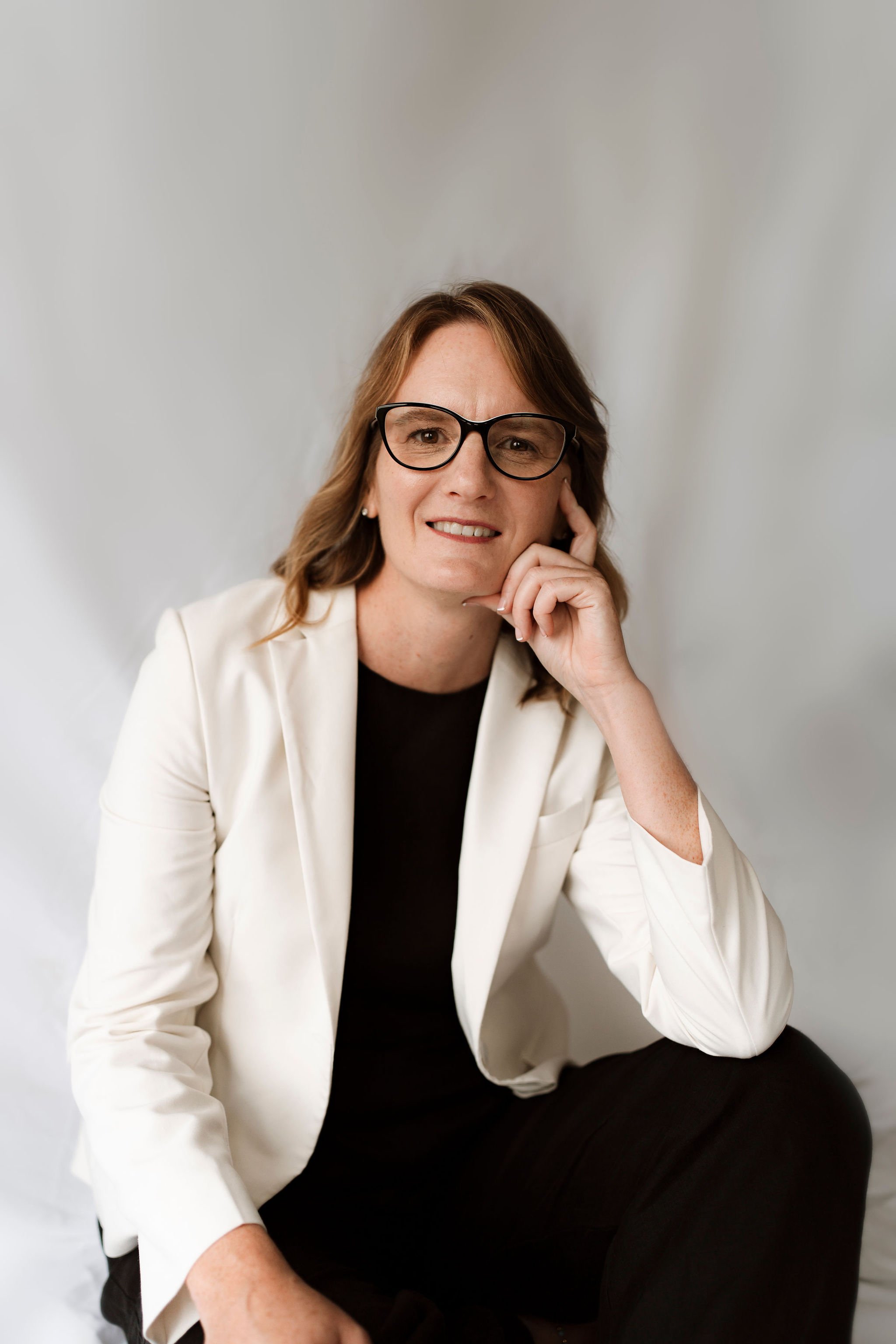 Woman wearing glasses and a white blazer seated with hand on chin, neutral background.
