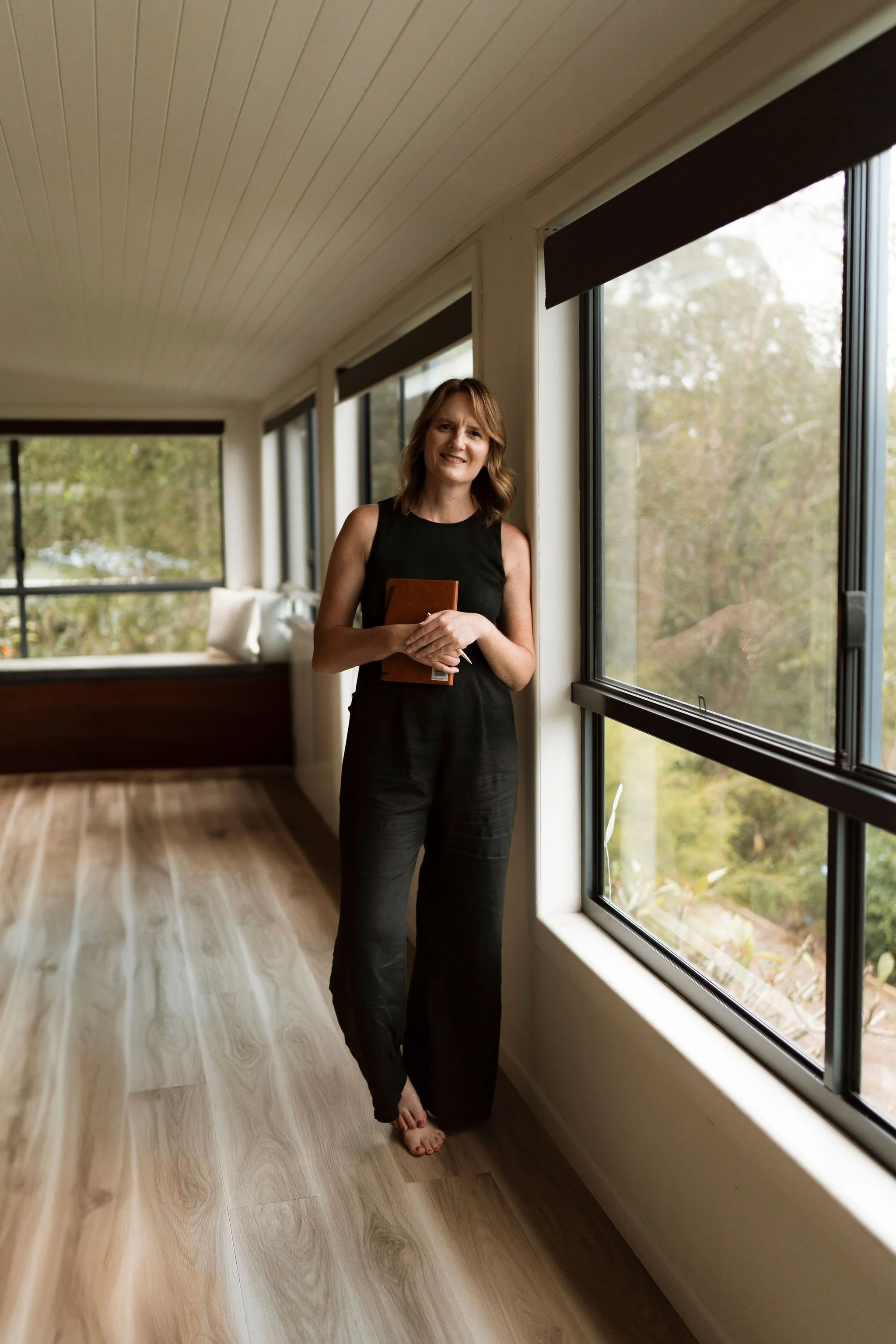 Woman standing barefoot in a sunlit room, holding a book, with large windows and outdoor garden view.