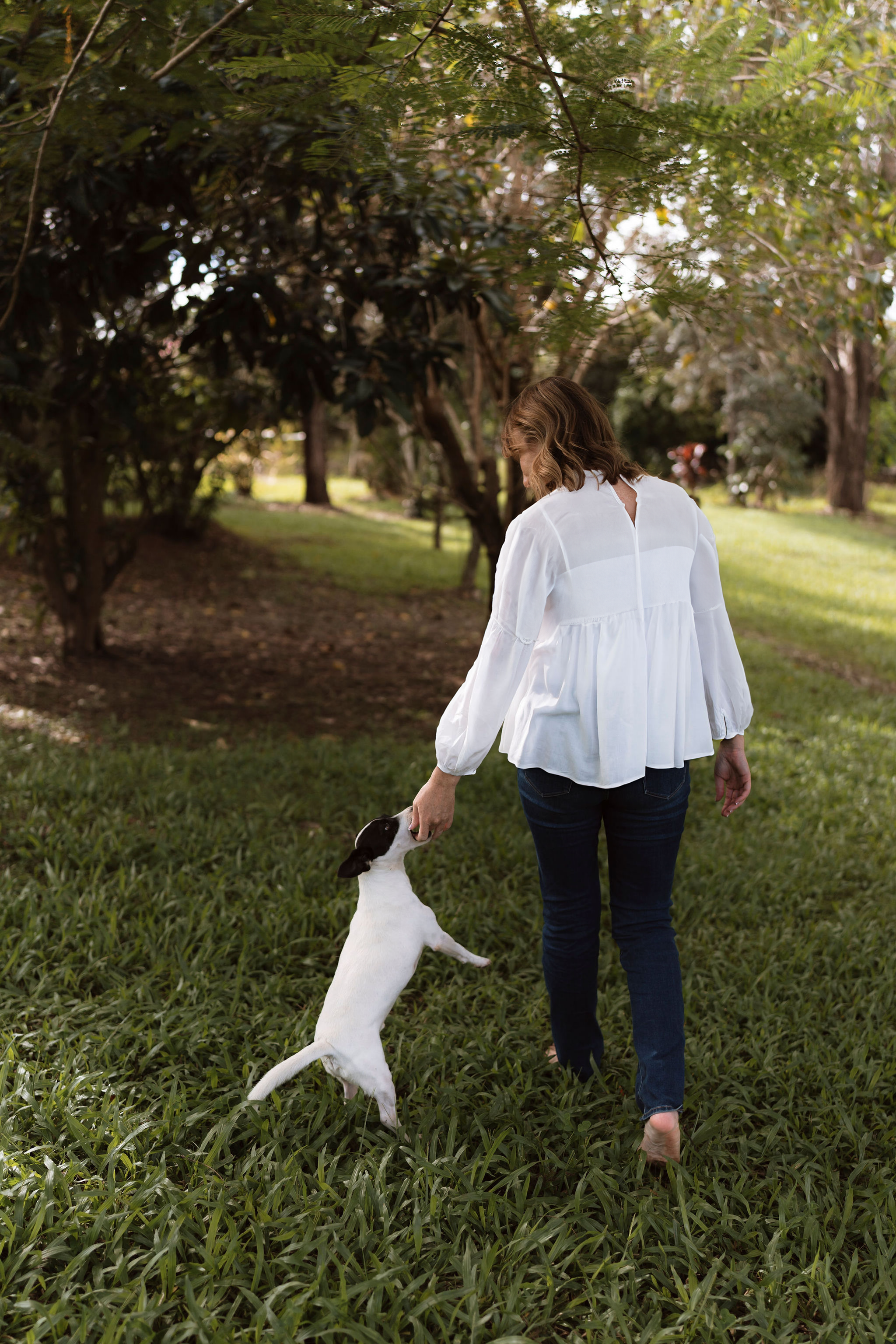 Woman in white blouse walking barefoot on grass with a jumping dog.