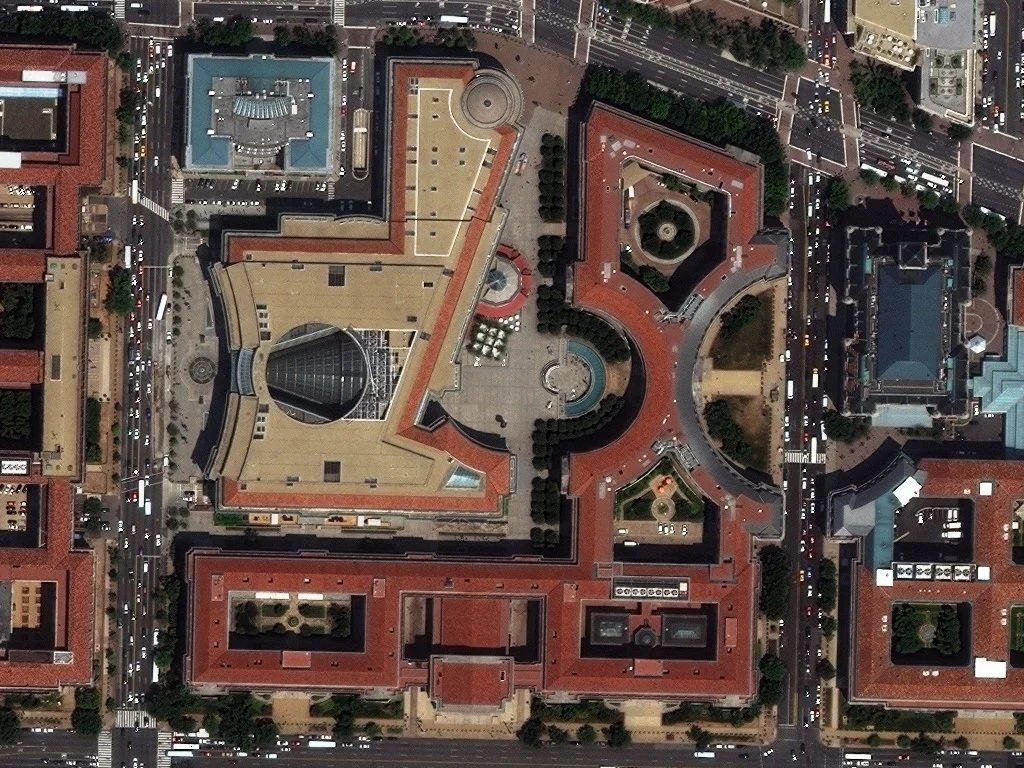 Aerial view of a complex city block with multiple large buildings, including a distinctive architecture with a glass dome, and surrounding streets with traffic.