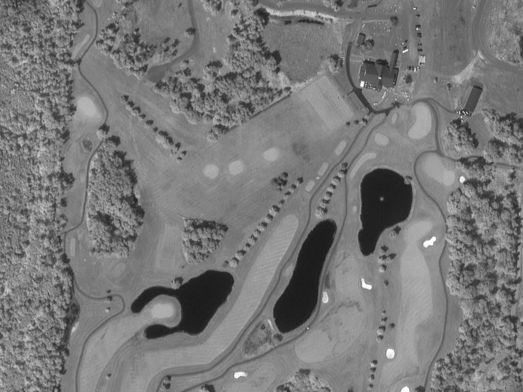 Aerial view of a golf course with fairways, water hazards, sand bunkers, and surrounding trees, with a clubhouse and parking lot at the top.