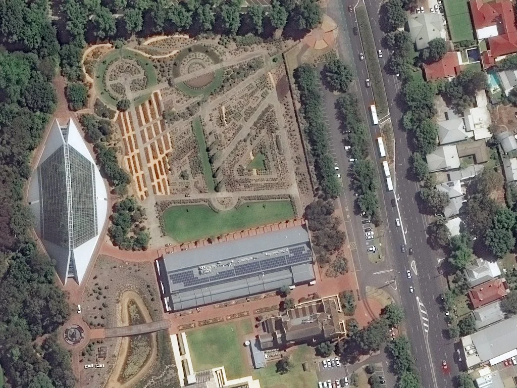 Aerial view of a park with pathways, gardens, and a greenhouse, next to a major street with cars and residential houses.