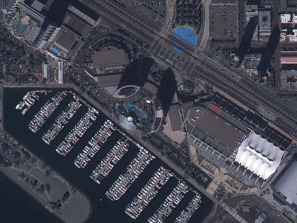 Aerial view of a marina with boats docked and adjacent buildings, including a large, curved white roof structure, and surrounding city infrastructure with roads and high-rise buildings.