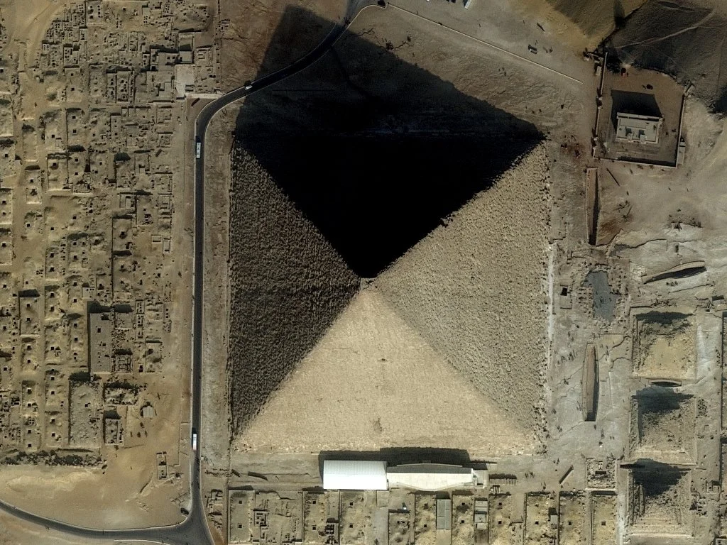 An aerial view of an ancient pyramid with a shadow cast on its side, surrounded by desert and archaeological ruins.