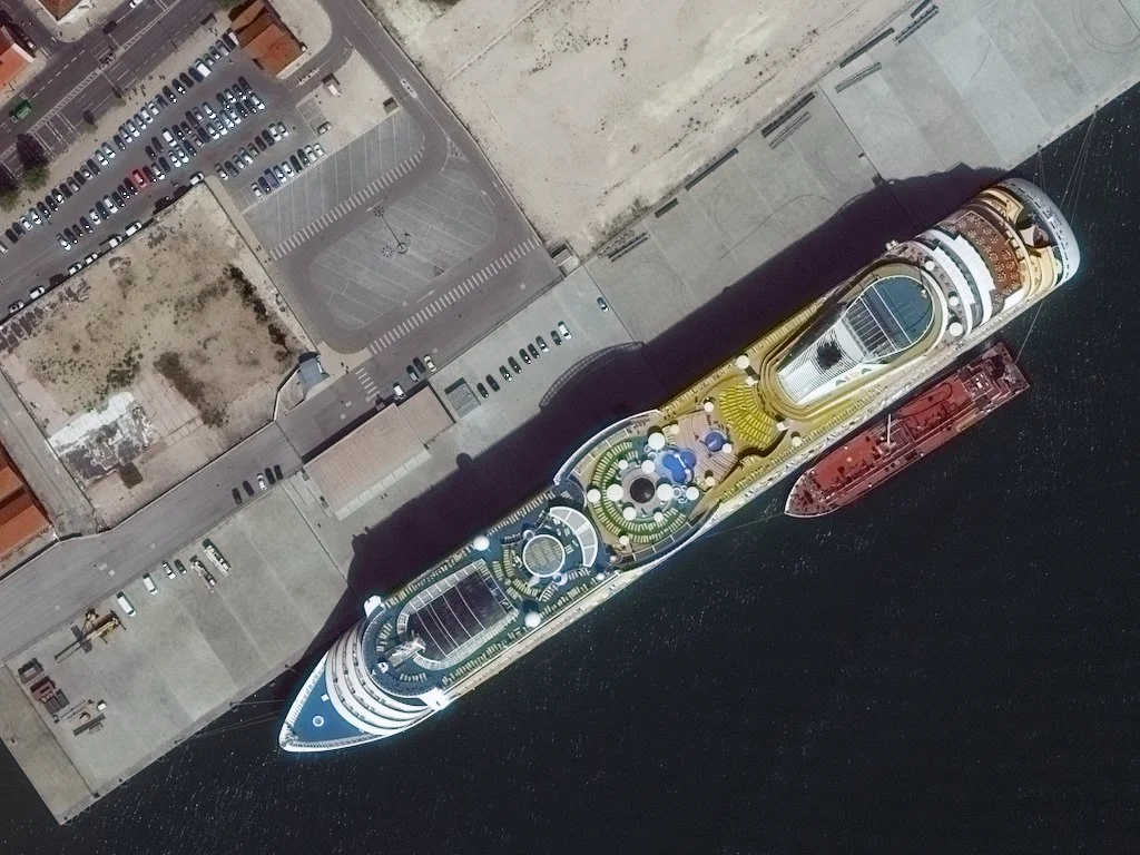 A cruise ship docked at the port, viewed from above, with parked cars and empty lots nearby.