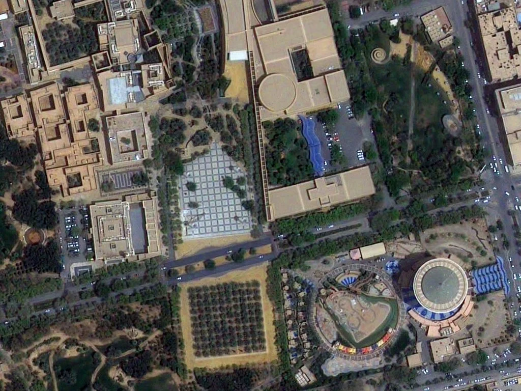 Aerial view of a cityscape showing buildings, parking lots, roads, green parks, and a large hotel or convention center with a distinctive round structure.