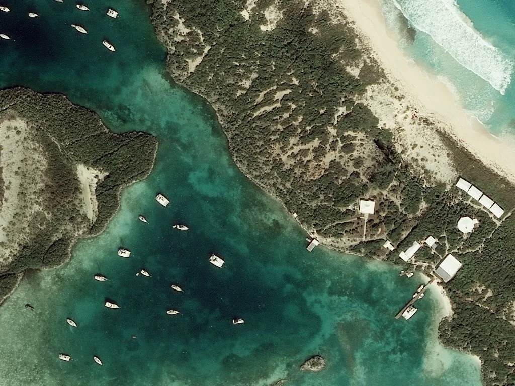 Aerial view of a coastal marina with boats, surrounded by green islands, with a sandy beach and waves from the ocean in the background.