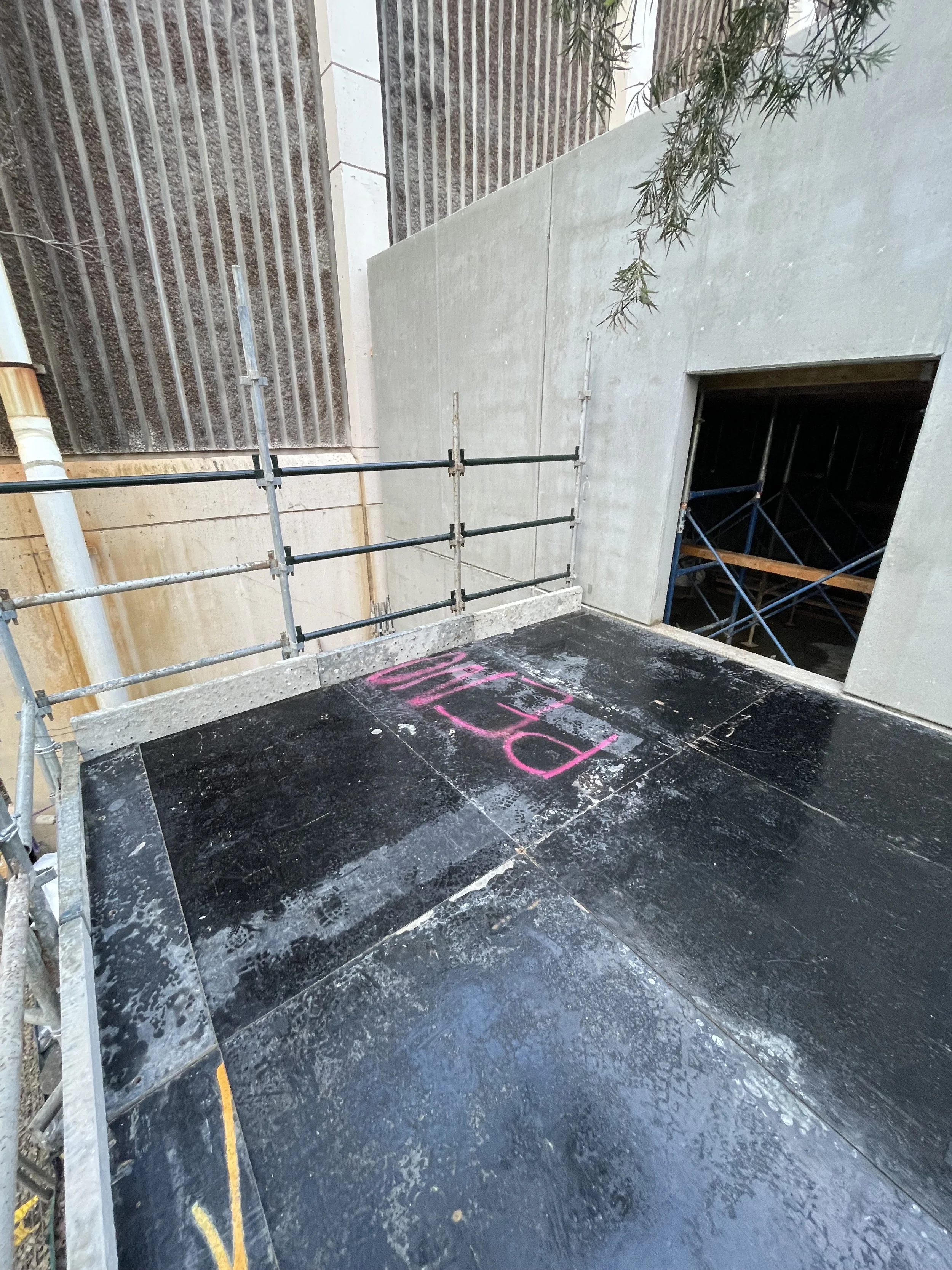 Construction site with wet black flooring, a concrete wall, and a small opening in the wall. Pink spray-painted number or word on the floor and scaffolding present.