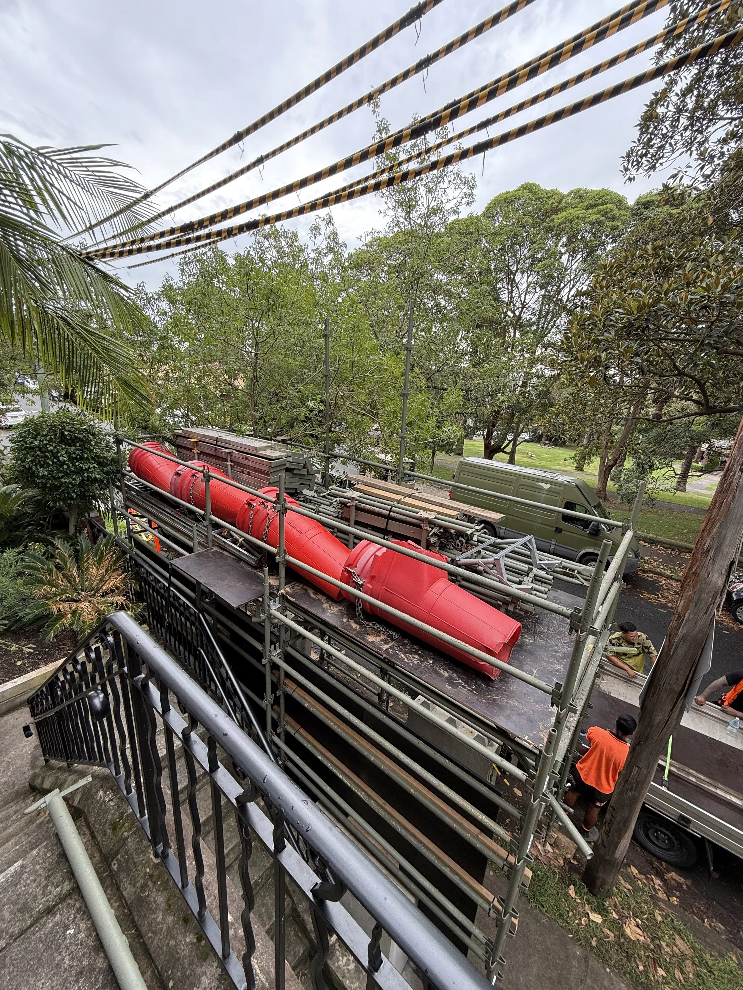 A truck with scaffolding and a large red pipe on its flatbed, parked on a residential street with trees and a few people around.