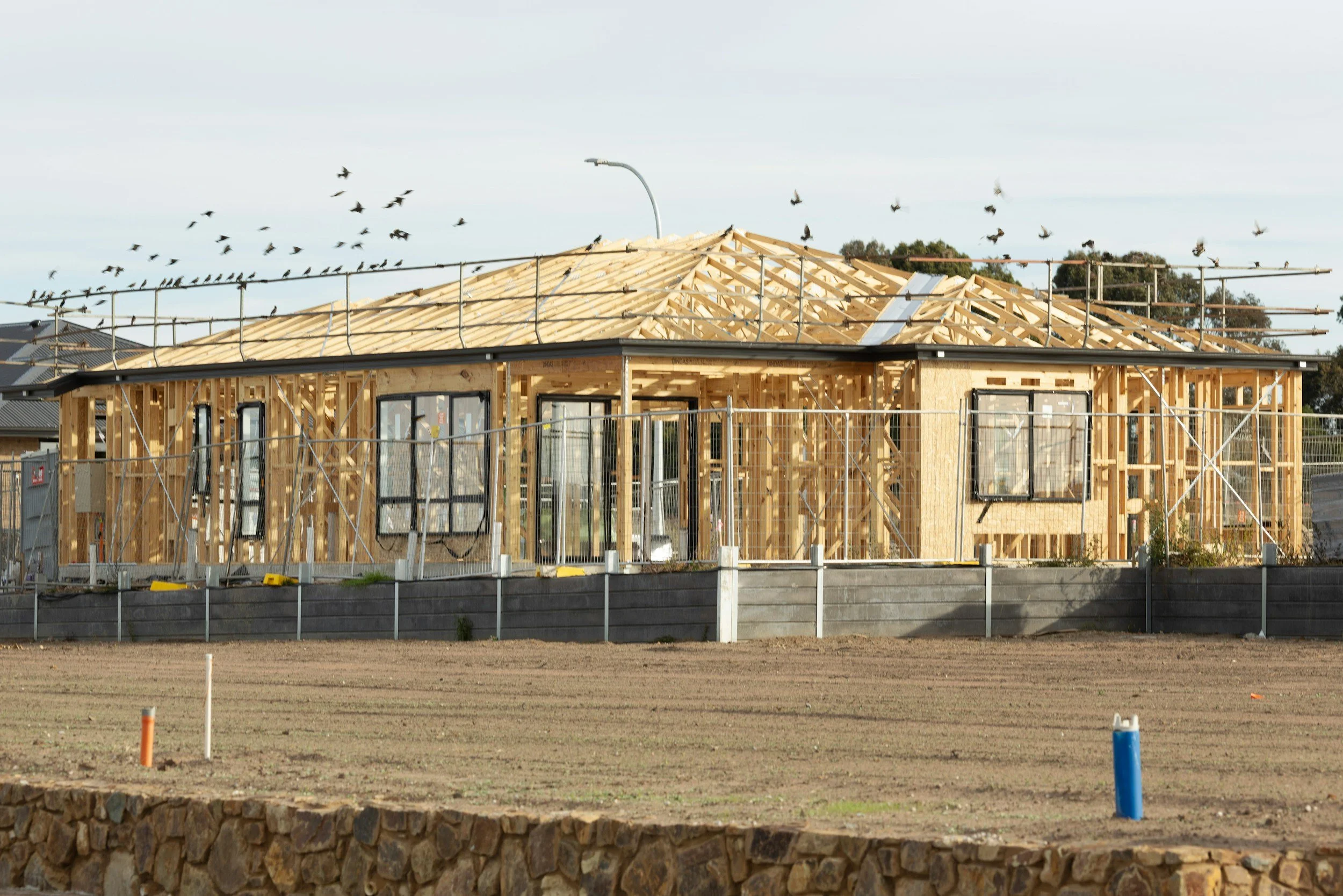 Wooden house under construction with framing in progress, surrounding a construction site with fencing and exposed soil, and birds flying in the sky.