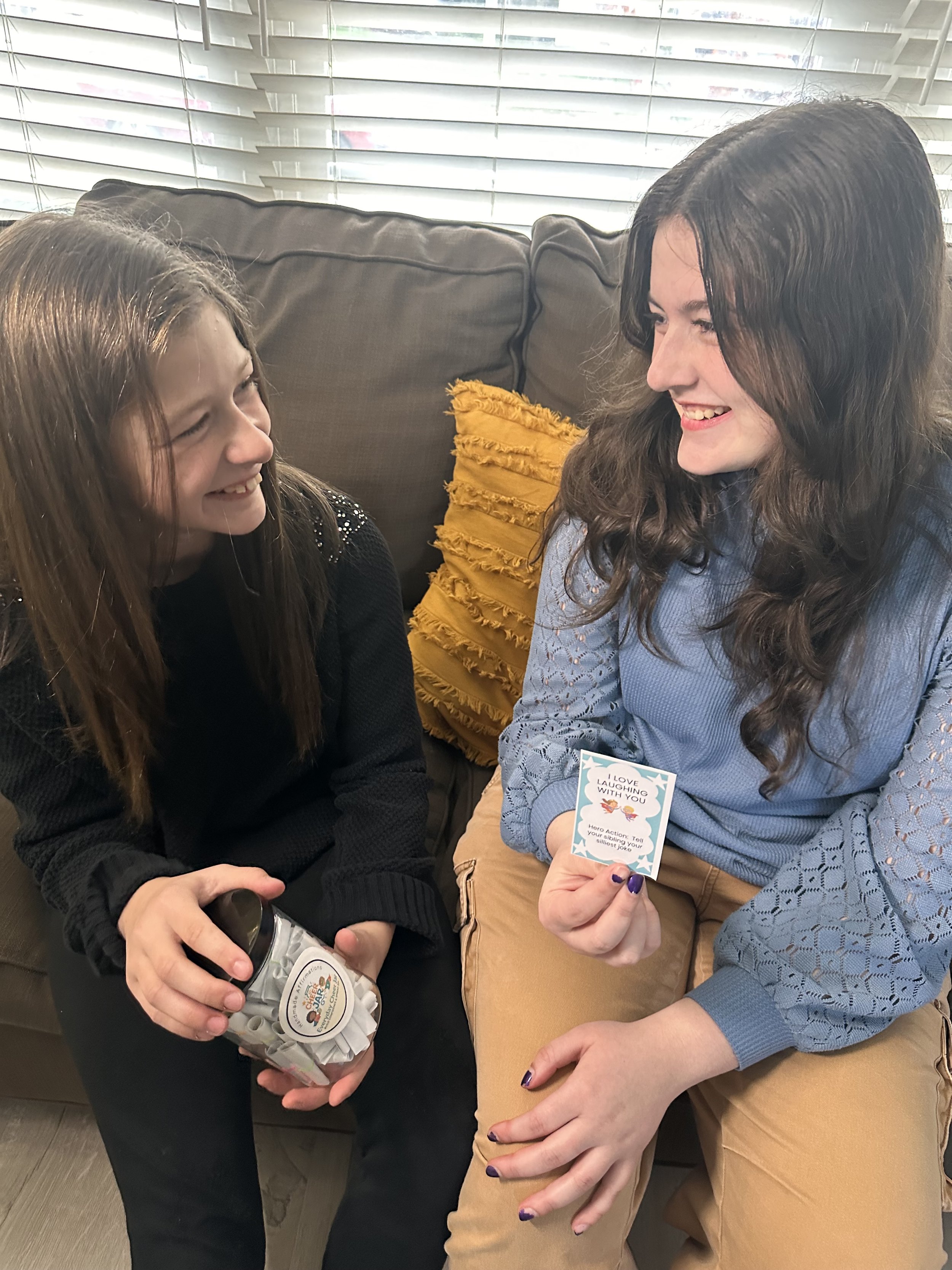 Two women sitting on a couch, smiling at each other. One is holding a container of cookies, and the other is holding a card that reads 'I love laughing with you'.