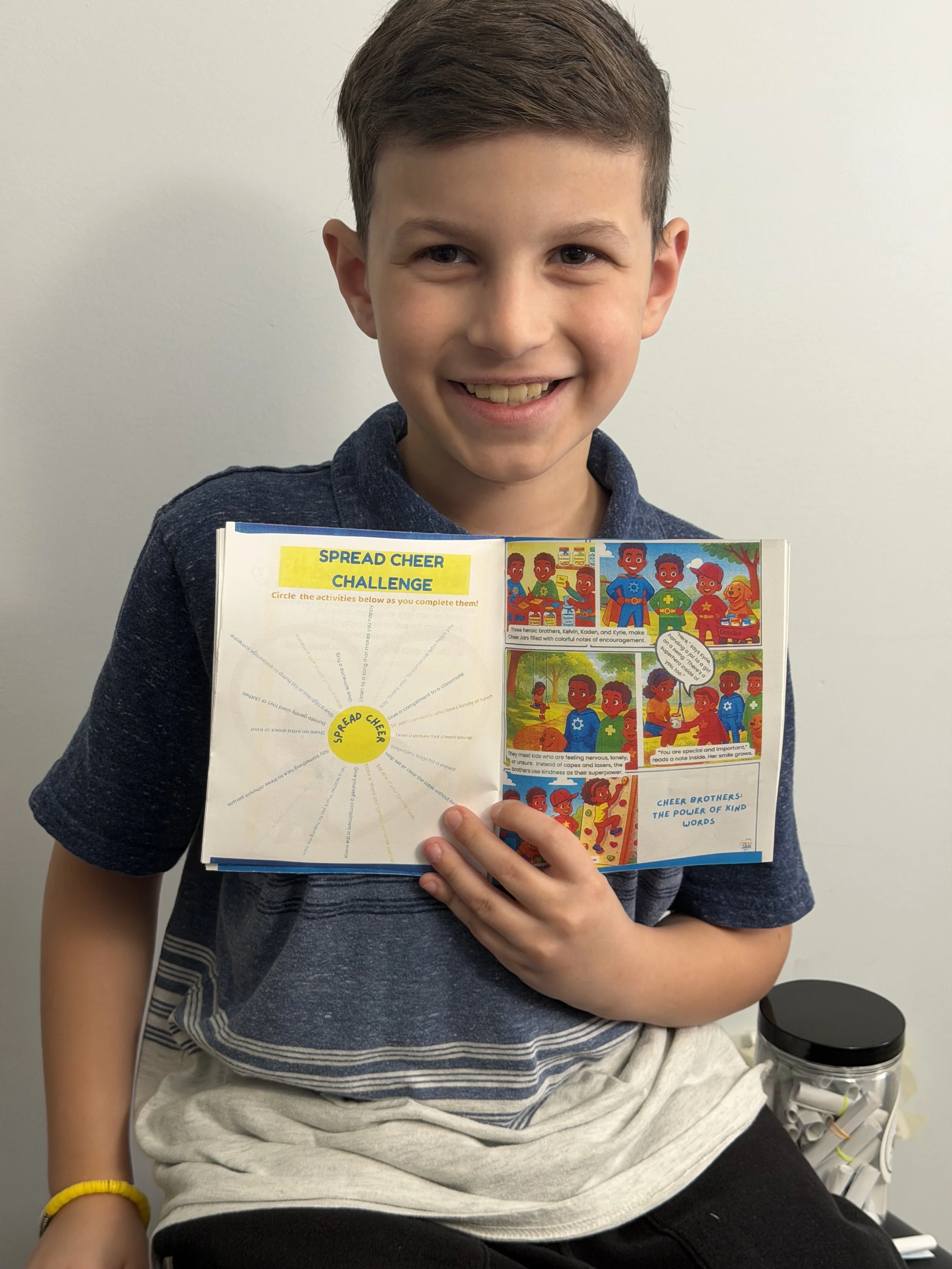 A young boy smiling and holding an open comic book titled 'Spread Cheer Challenge'. The comic features colorful illustrations of children encouraging kindness and cheer among brothers.