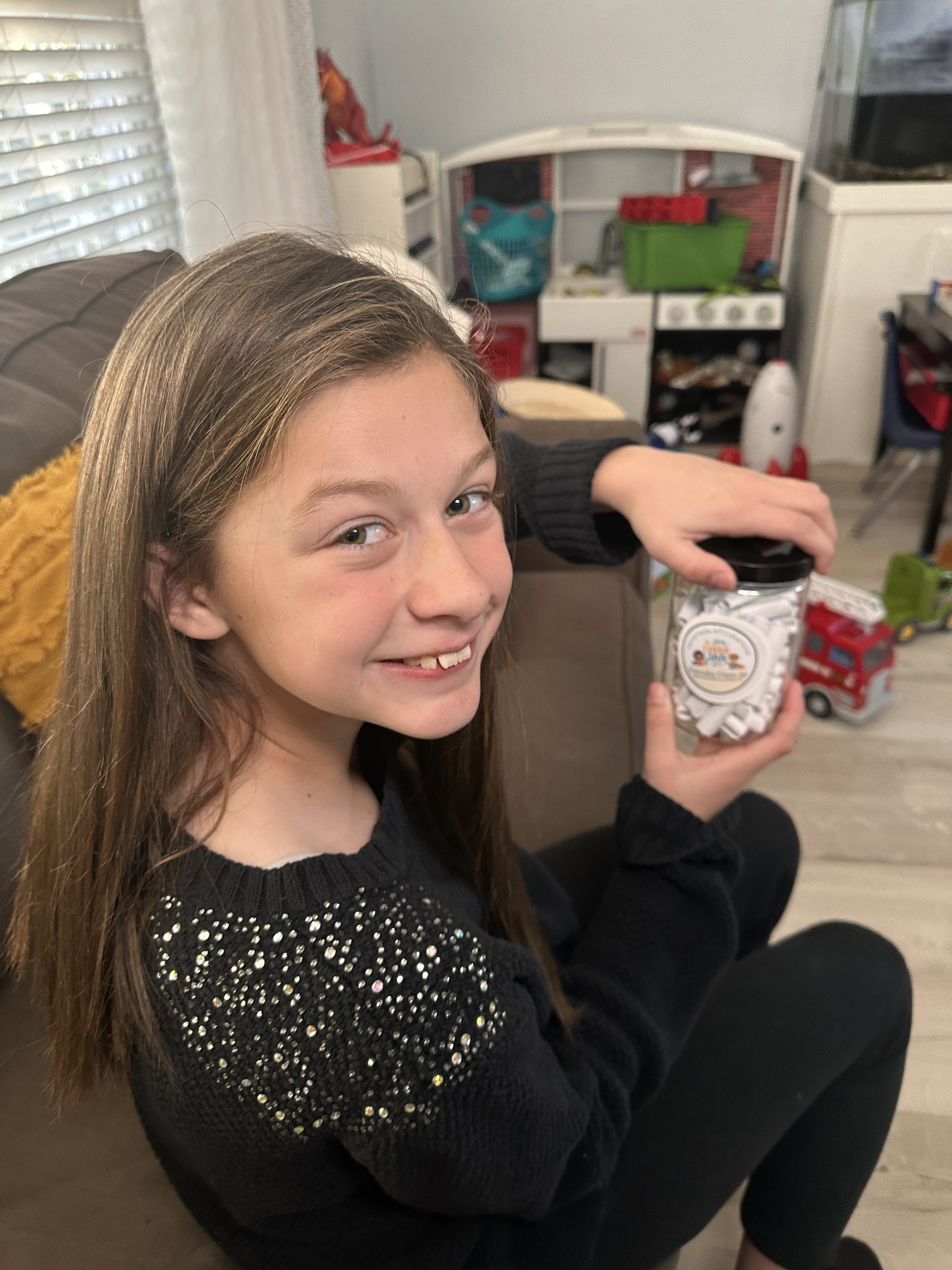 A smiling girl with long hair holding a jar of white candies indoors, with a background of a toy kitchen and toys on the floor.