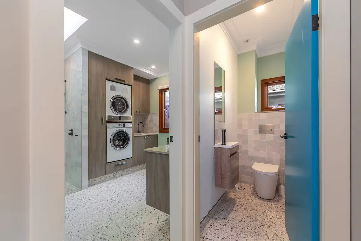 View of a laundry room with stacked washer and dryer, with a bathroom featuring a toilet, small vanity with mirror, and a window to the right.