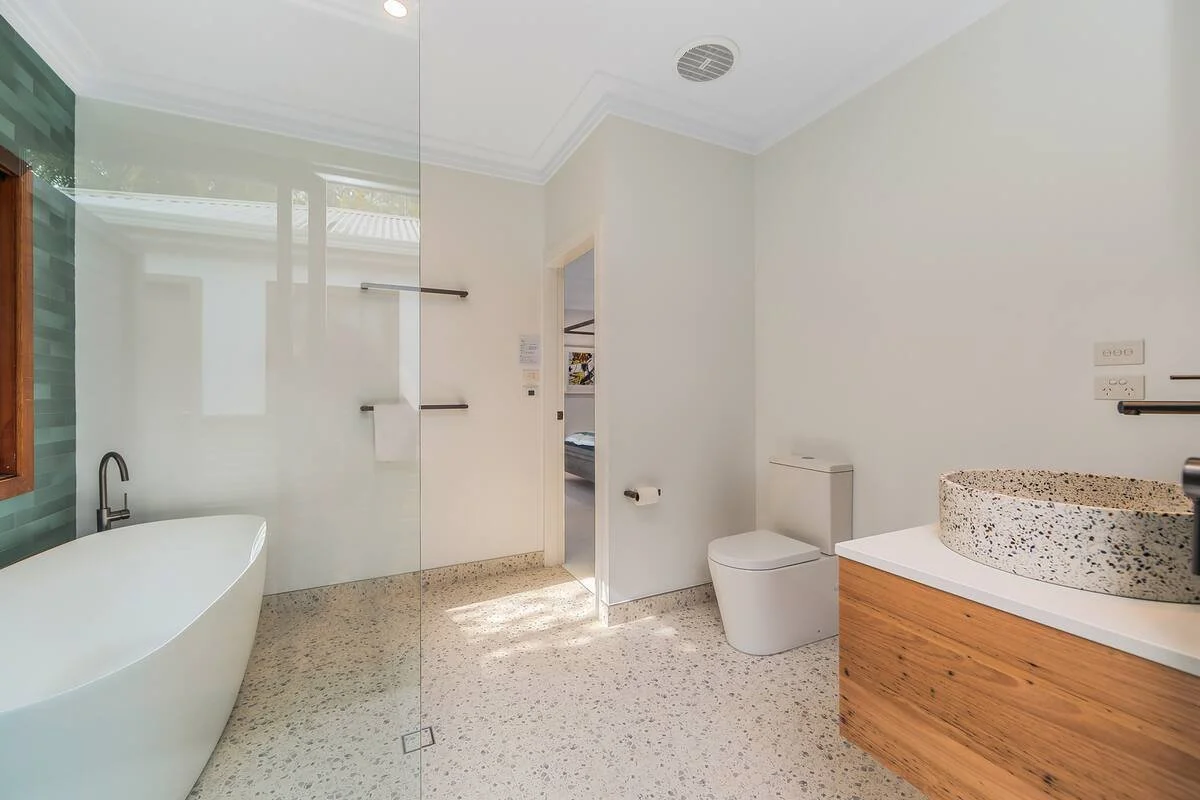 Modern bathroom with a clawfoot bathtub, walk-in shower, white toilet, wooden vanity with a speckled stone vessel sink, and a window with greenery outside.