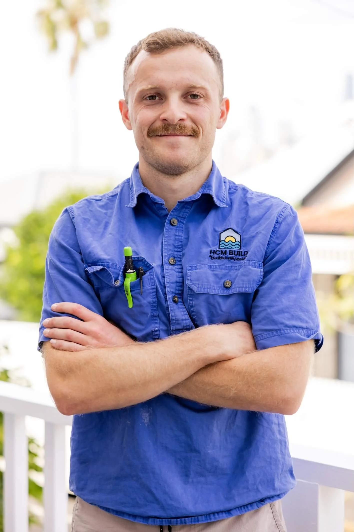 A man with a mustache wearing a blue work shirt with a logo, standing outdoors with arms crossed, smiling.