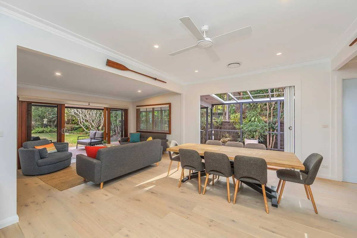 Open living and dining area with wooden floors, gray sofas, yellow and teal cushions, a wooden dining table with eight chairs, sliding glass doors leading to a garden, and a ceiling fan.