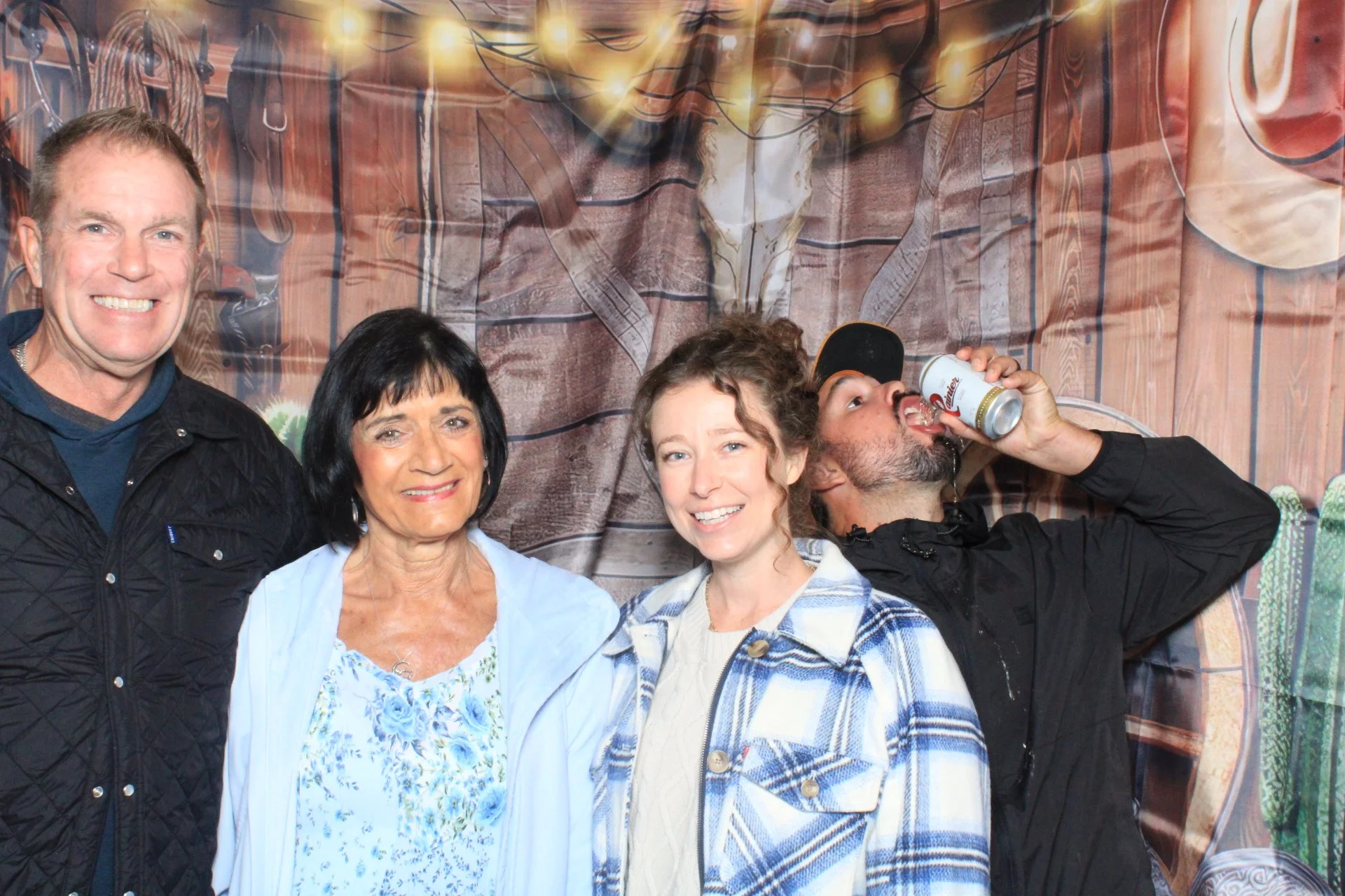 Four people standing in front of a Western-themed backdrop. The group includes two women and two men, with one man drinking from a can.